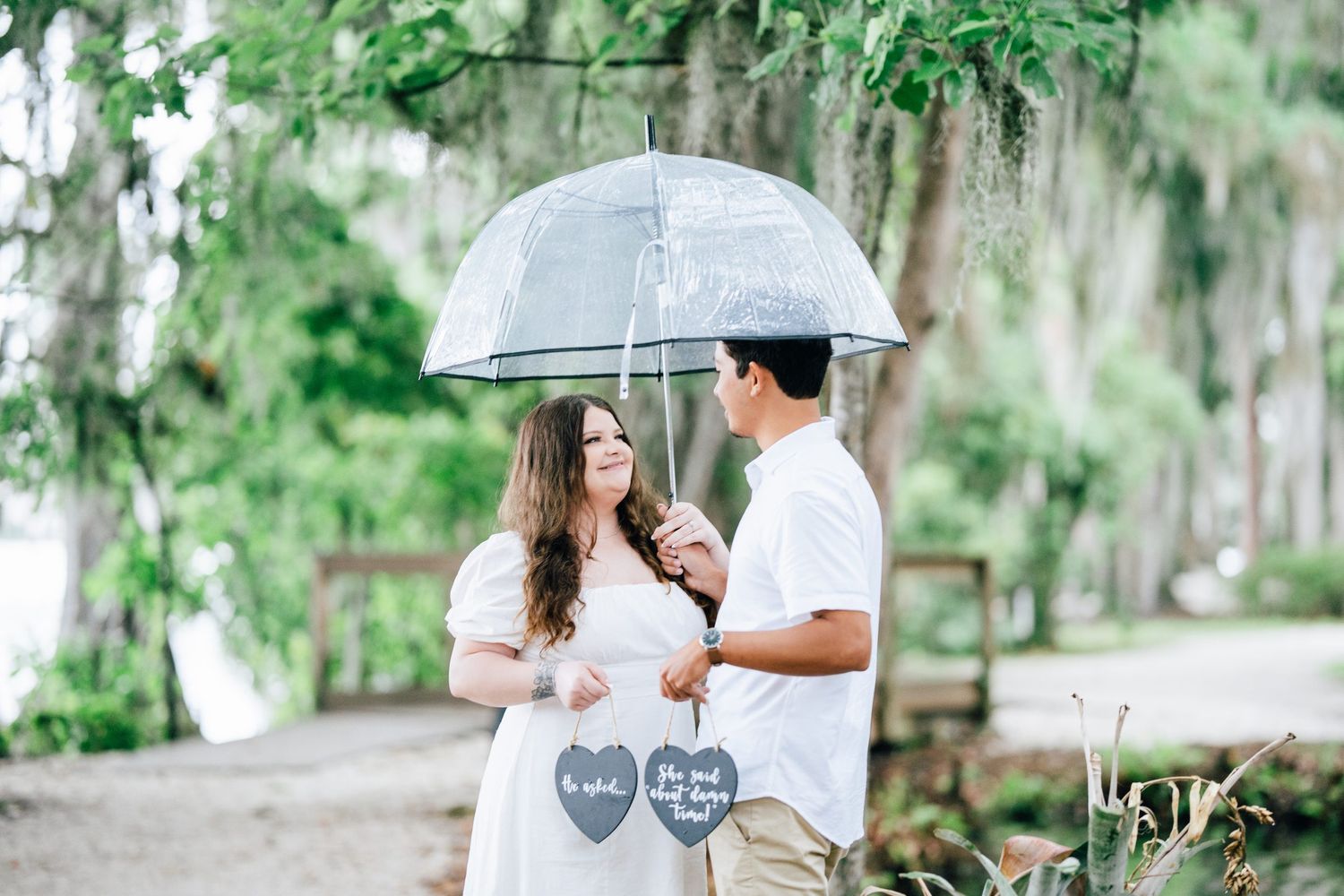 Couple sharing a clear umbrella in a lush garden, holding heart-shaped signs, dressed in white for an engagement photo.