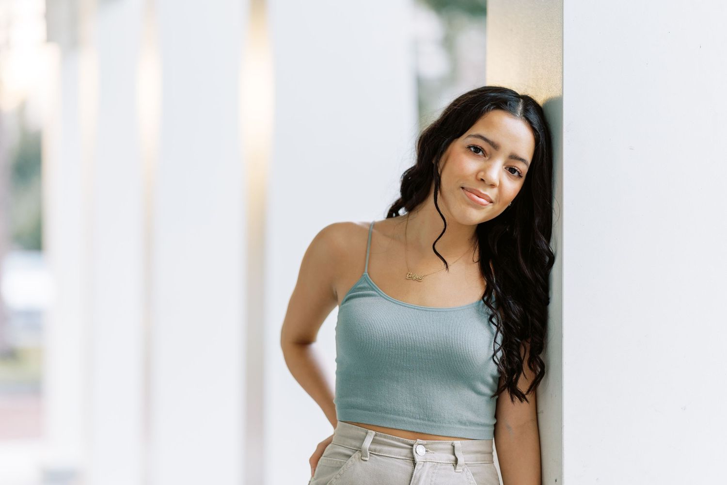 A woman in sage green tank top and khaki pants stands against a white wall with soft natural lighting.