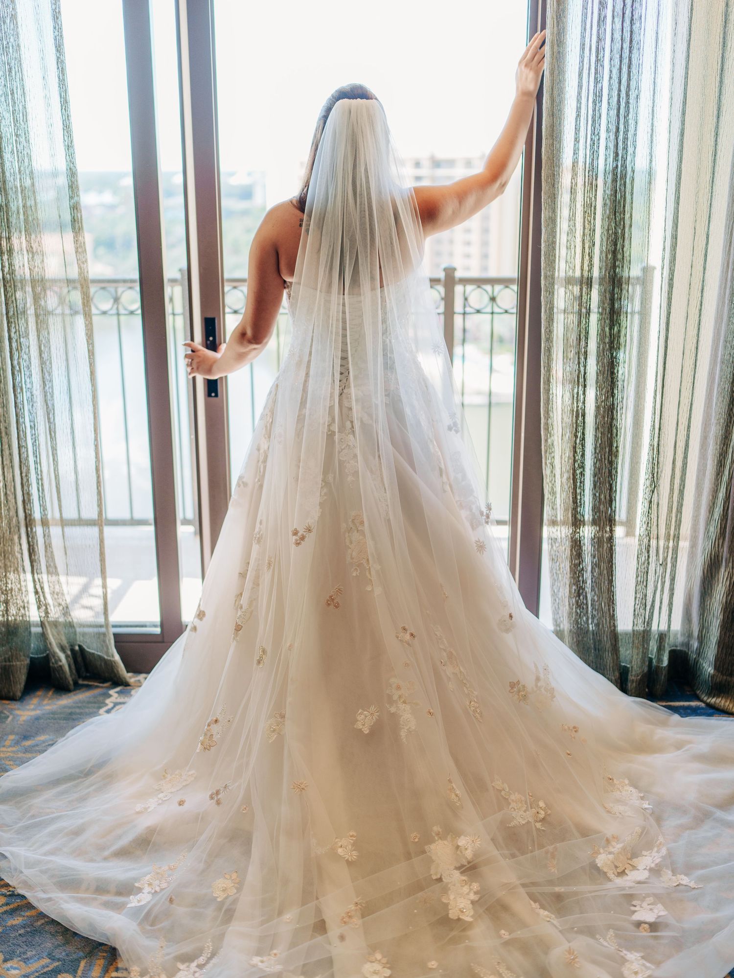 A bride in a flowing white wedding gown with floral appliques and cathedral length veil stands by sheer curtained windows.
