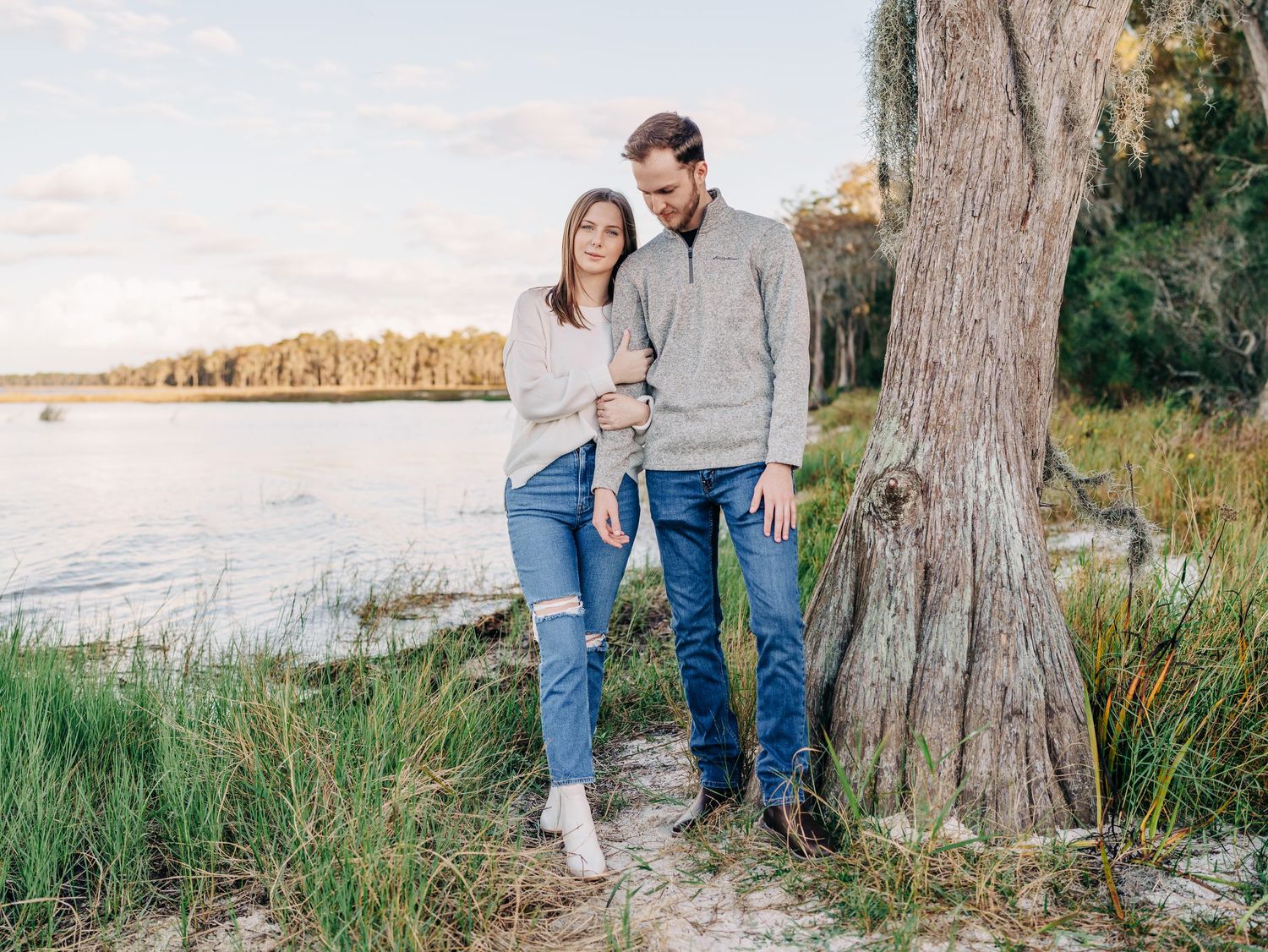 Couple embracing near a lakeside tree on a sandy shore, wearing casual jeans and sweaters in a serene outdoor setting.