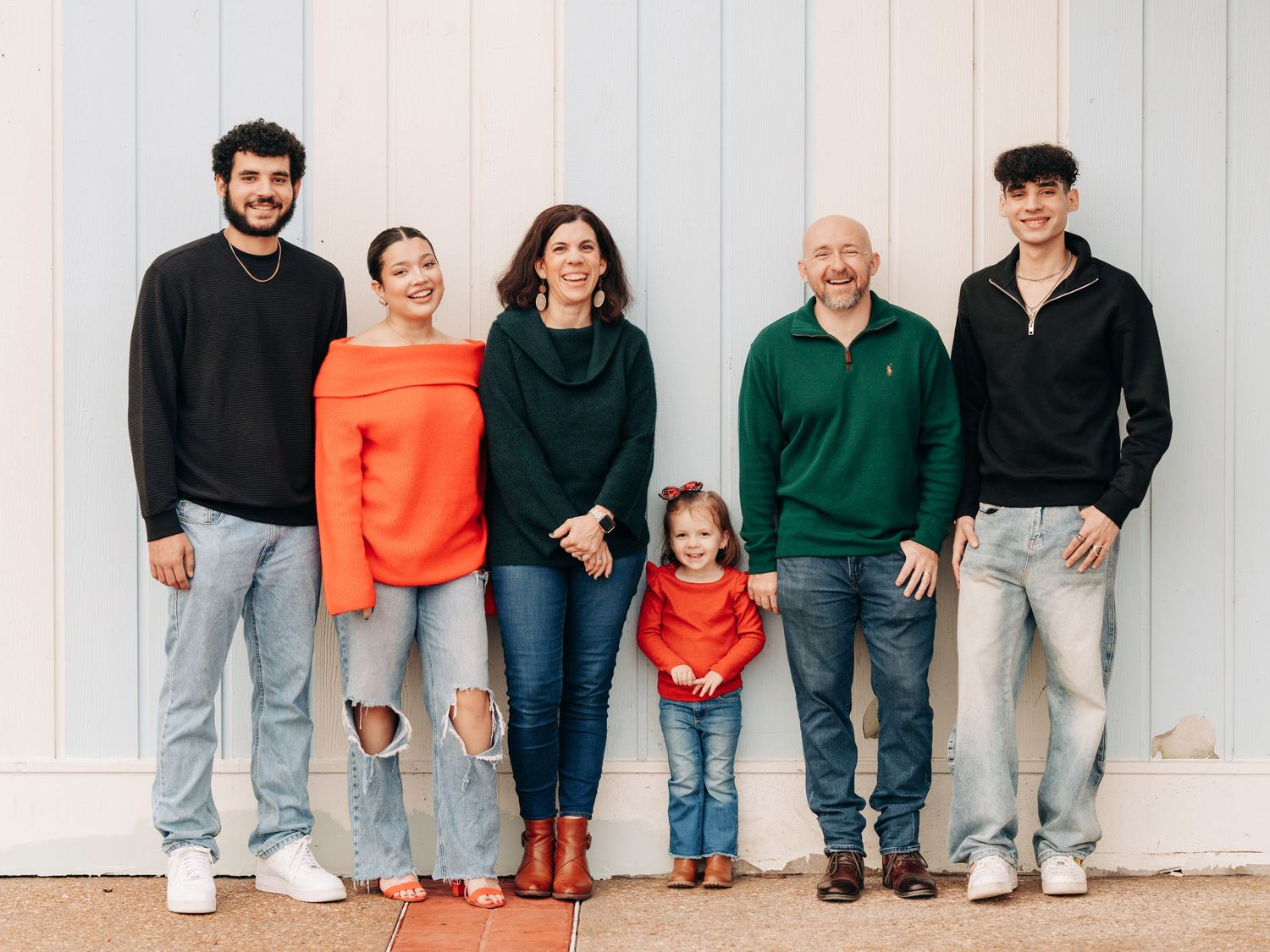 A group of five people wearing casual clothing and denim stand together against a white wall outdoors.