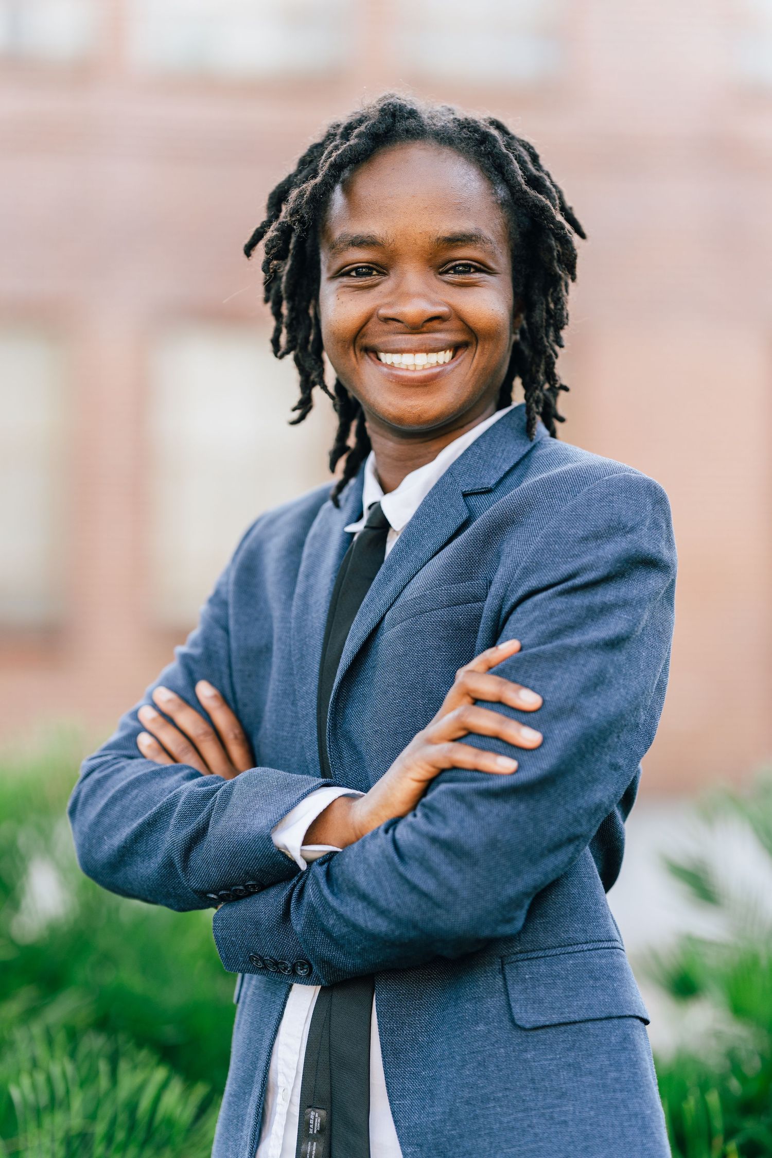Professional wearing a navy business suit and tie stands with arms crossed while smiling outdoors near a building.