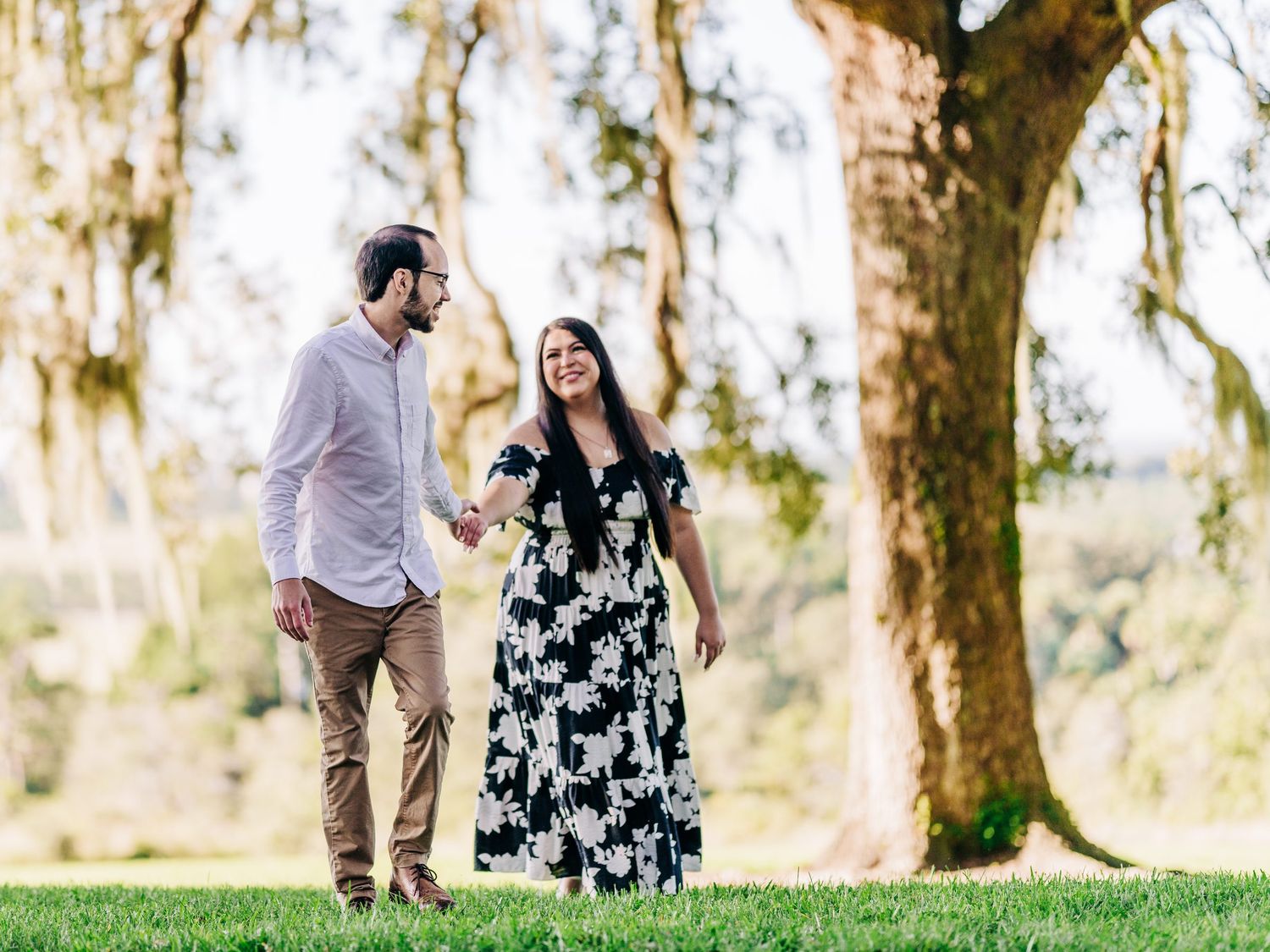 Couple walking hand in hand on green grass beneath large oak trees in a sunny park, man in pink shirt, woman in floral dress.