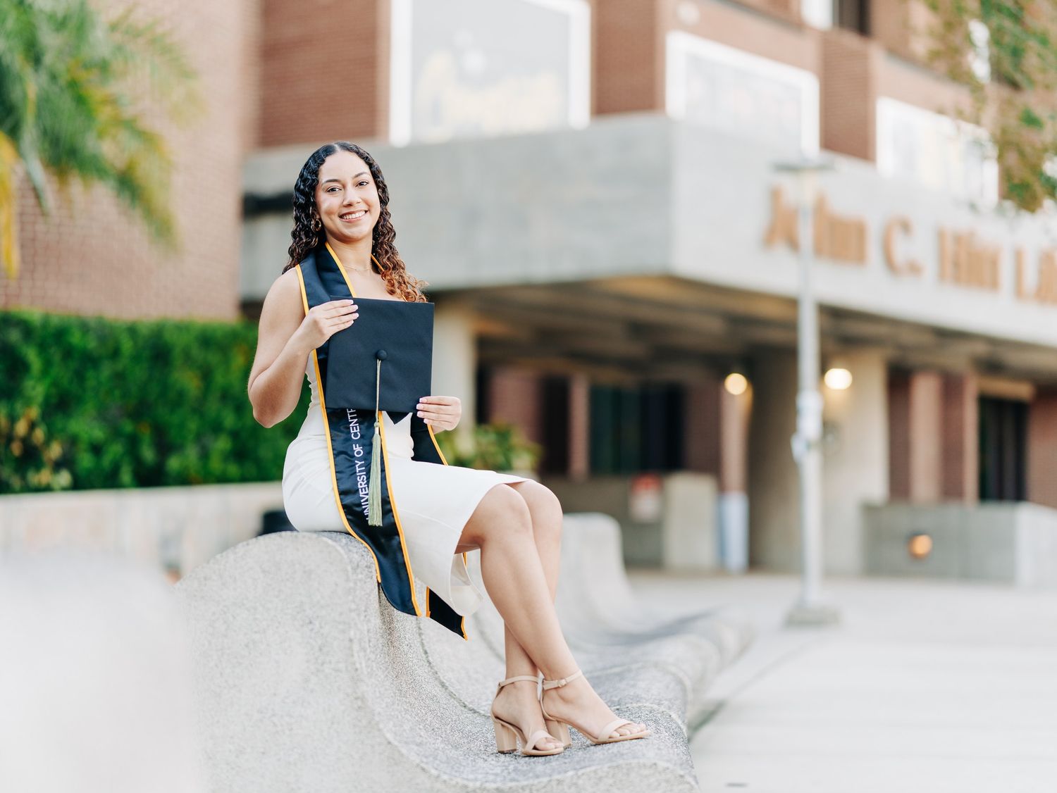 Smiling female graduate in white dress holding cap and blue sash, seated outdoors near university building on sunny day.