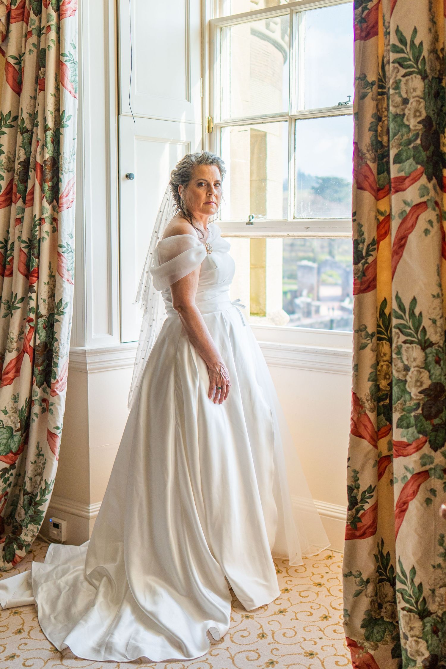 Elegant bride in flowing white satin wedding gown poses by window with floral curtains in natural light.