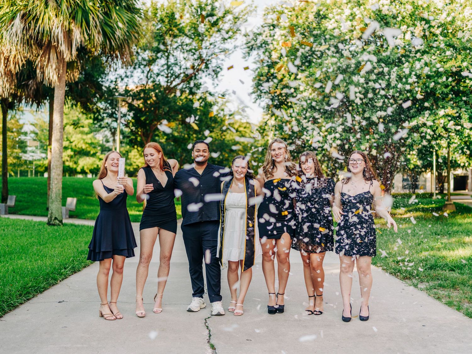 A group wearing cocktail dresses and formal attire pose together on a garden path surrounded by palm trees and greenery.