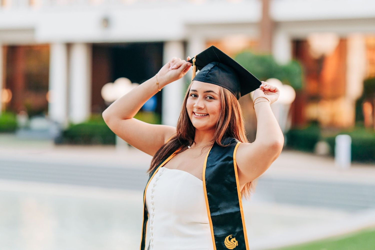Smiling female graduate in black cap and stole posing outdoors at university campus graduation ceremony.