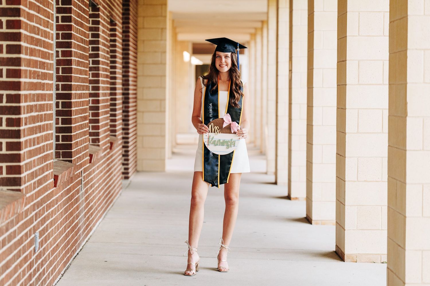Smiling graduate in cap and gown holds diploma in a brick building hallway, wearing white dress and heels.
