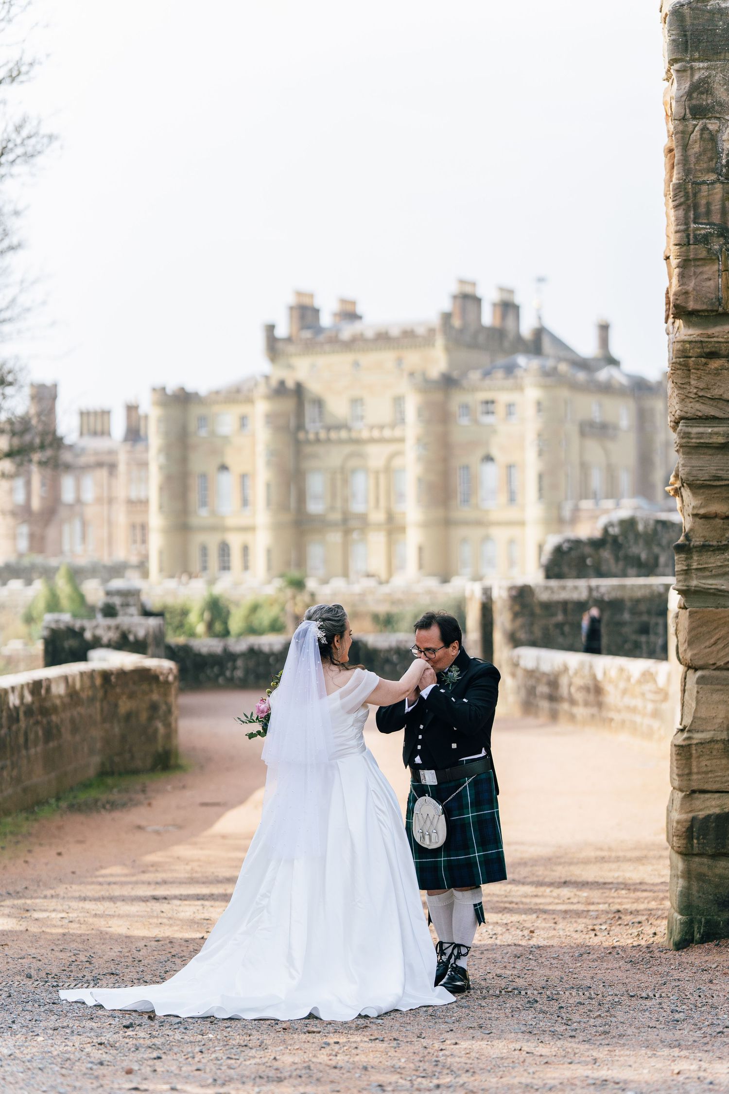 A couple shares a romantic moment in traditional wedding attire with a Scottish kilt against a grand castle backdrop.