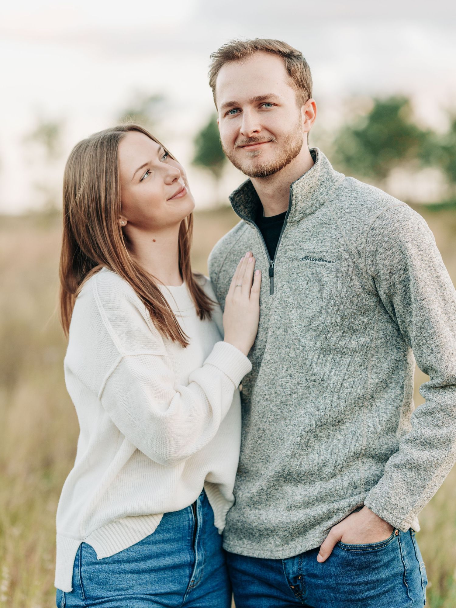 A couple wearing coordinating white and gray sweaters pose together in a natural outdoor setting at sunset.