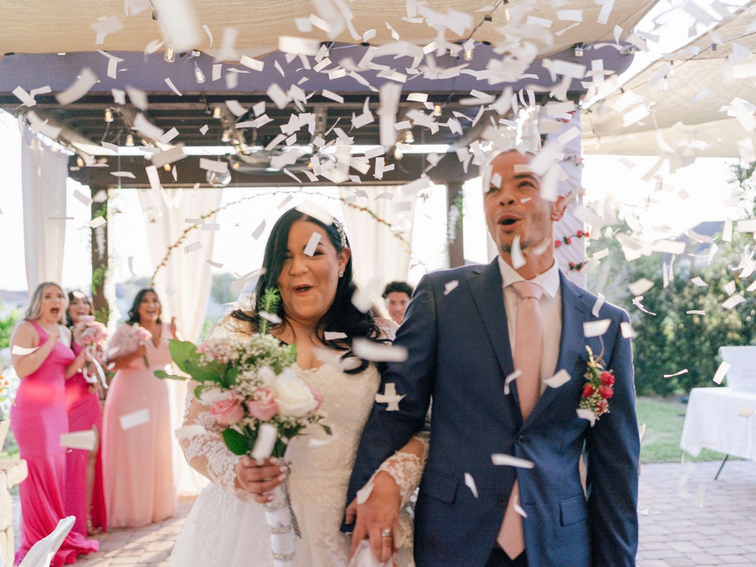 A newlywed couple celebrates joyfully under falling confetti while walking down an outdoor aisle with guests in pink dresses.