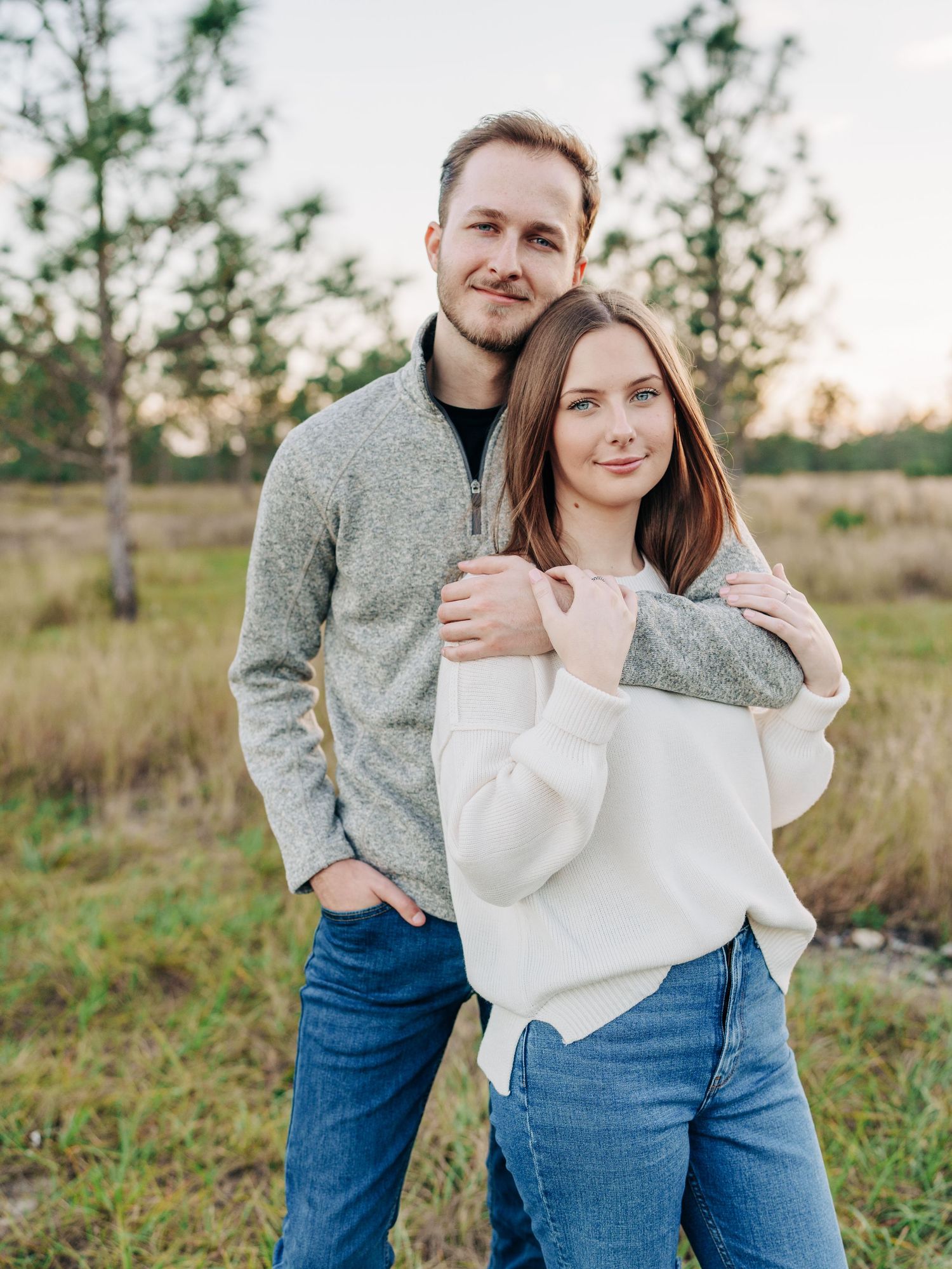A couple in casual sweaters embrace outdoors in a natural field setting during golden hour.