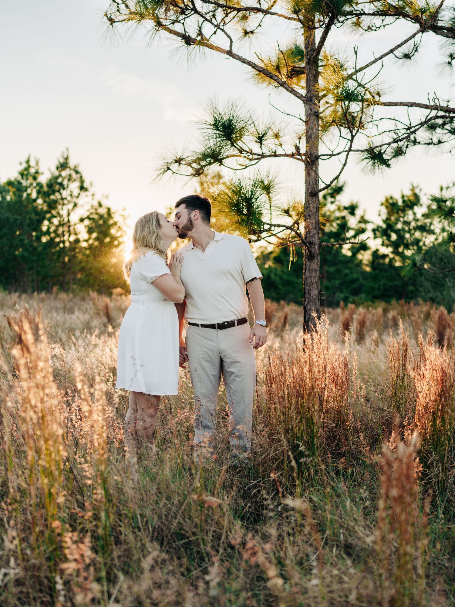 Couple embraces in golden light among tall grass and pine trees at sunset, wearing white and neutral summer outfits.