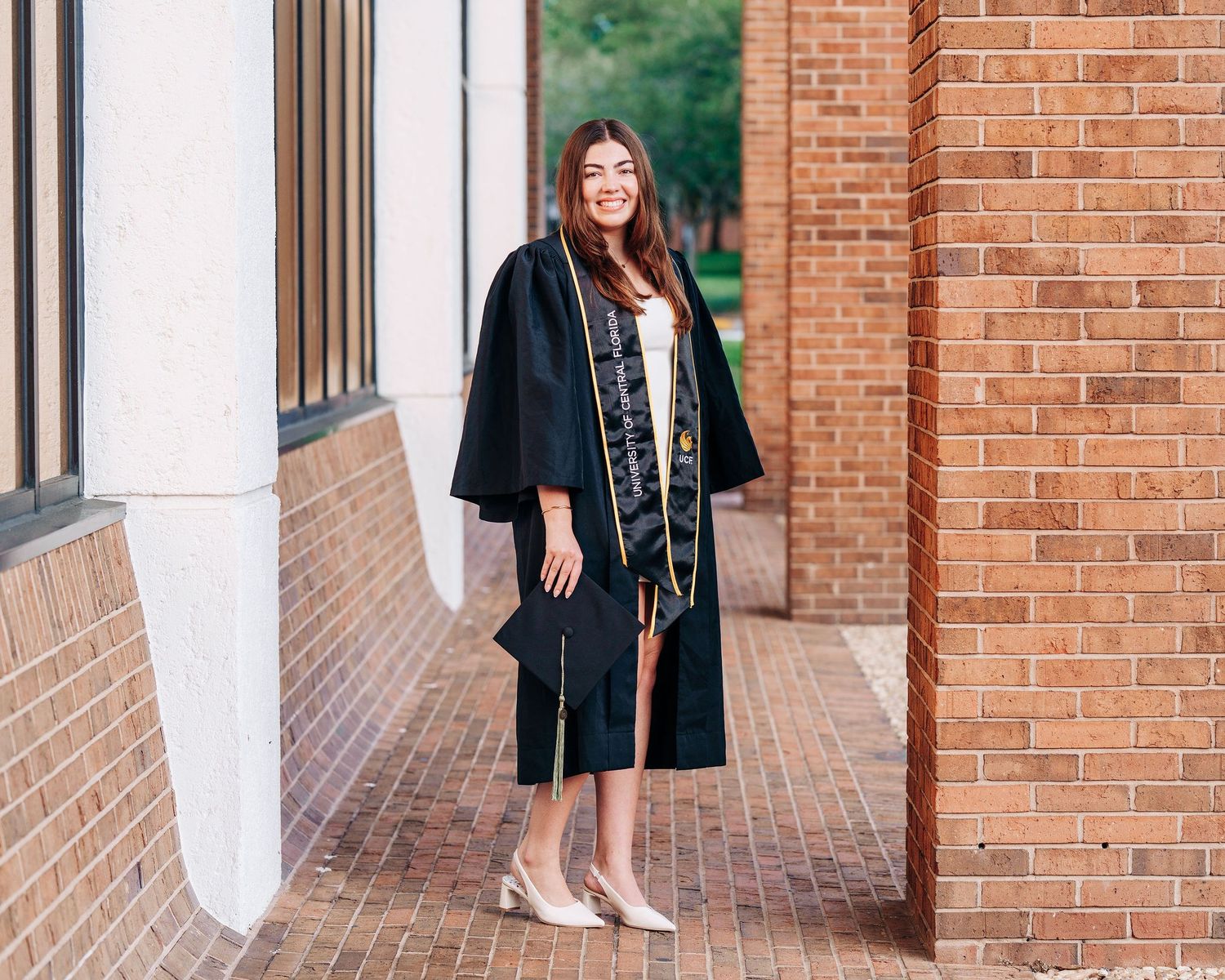 Young woman in black graduation cap and gown smiling outdoors near brick building columns.