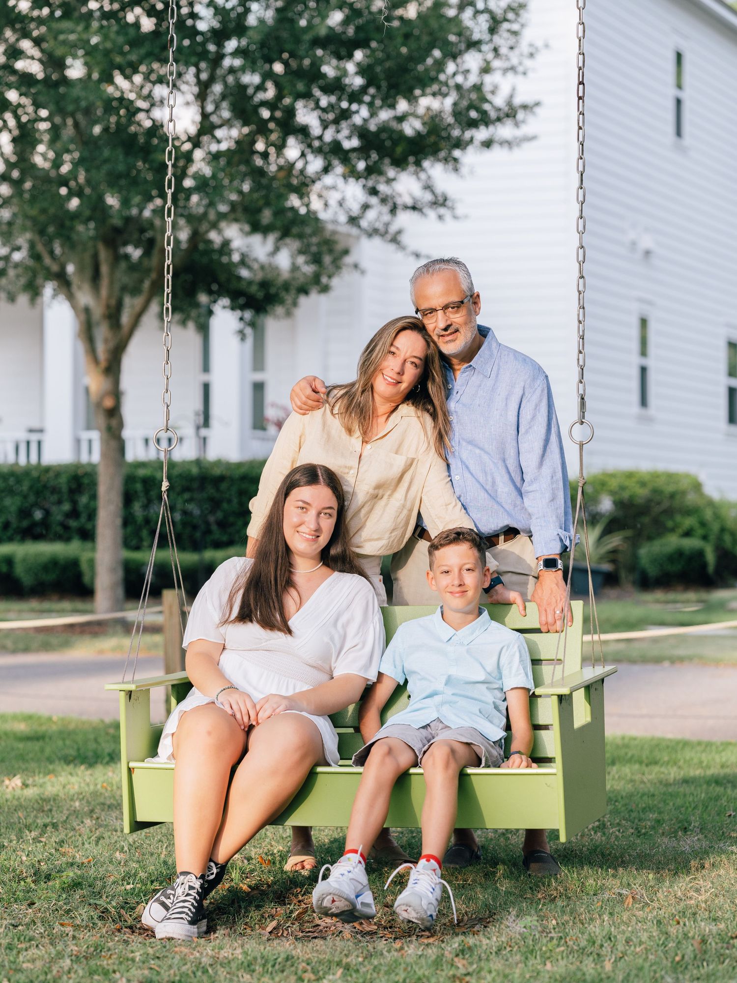 A family sits together on a lime green porch swing in front of a white house on a sunny day.