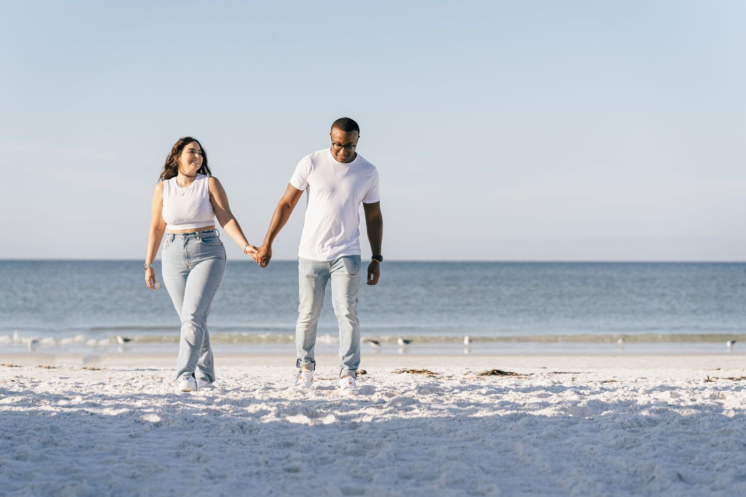 Couple dressed in white walking hand in hand along a sunny beach with ocean view in the background.
