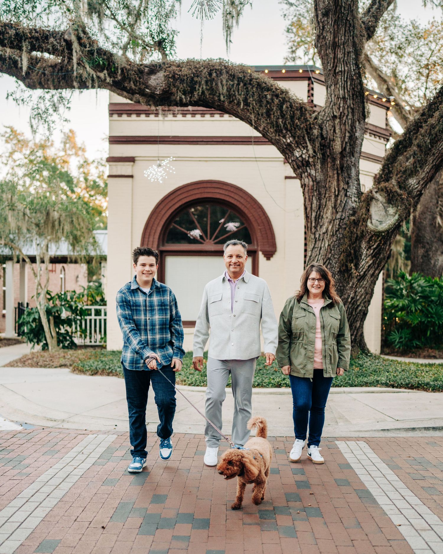 A family standing with their dog on a brick walkway in front of an arched entrance to a Spanish-style building.