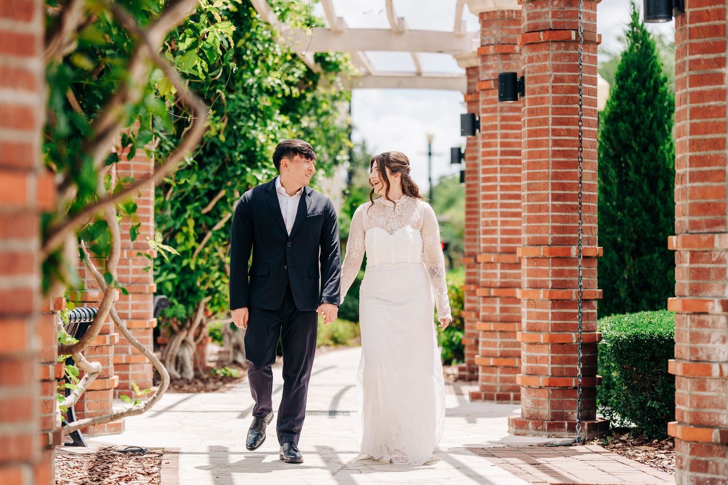 Bride and groom walk hand-in-hand through a brick archway garden path on their wedding day.