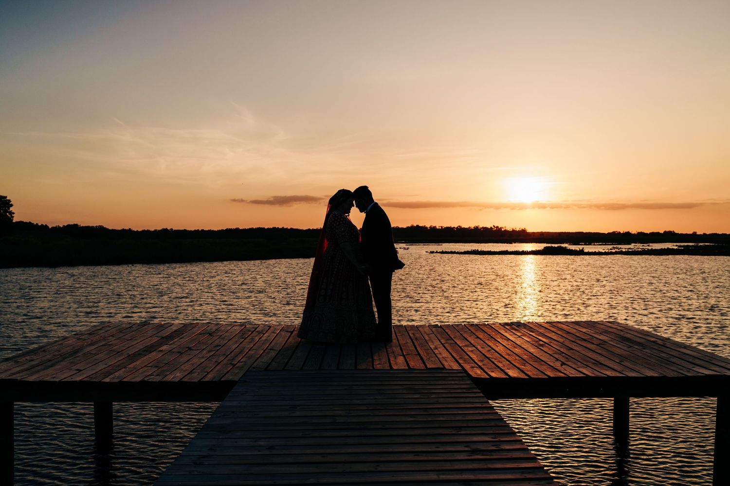 Silhouette of couple embracing on pier at sunset over tranquil Lavender on the Lake wedding venue lake creating a romantic waterfront scene.