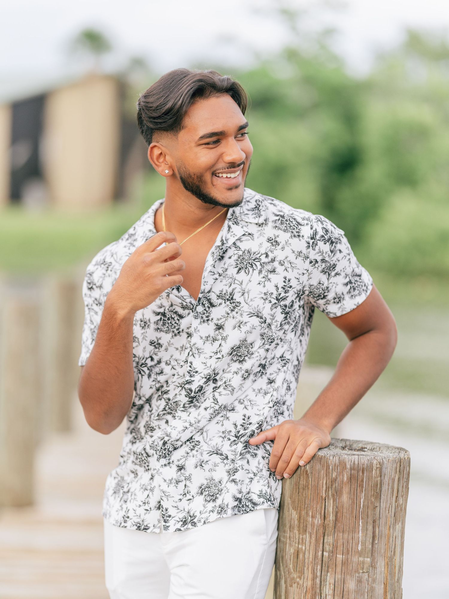 A person wearing a white and gray floral print shirt leans against a wooden post while smiling outdoors.