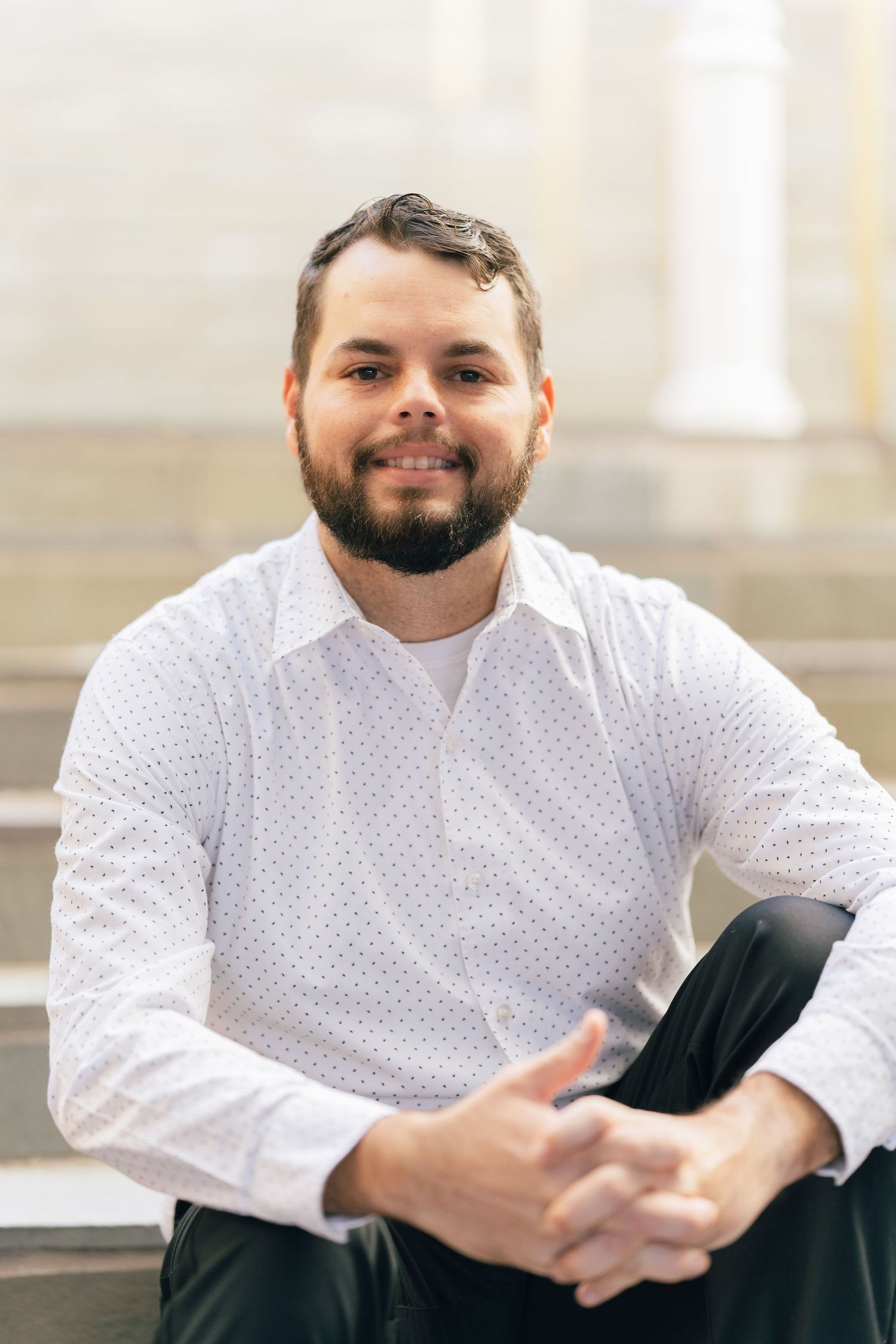 Professional portrait of someone in a white polka dot shirt sitting on outdoor steps with a relaxed pose.