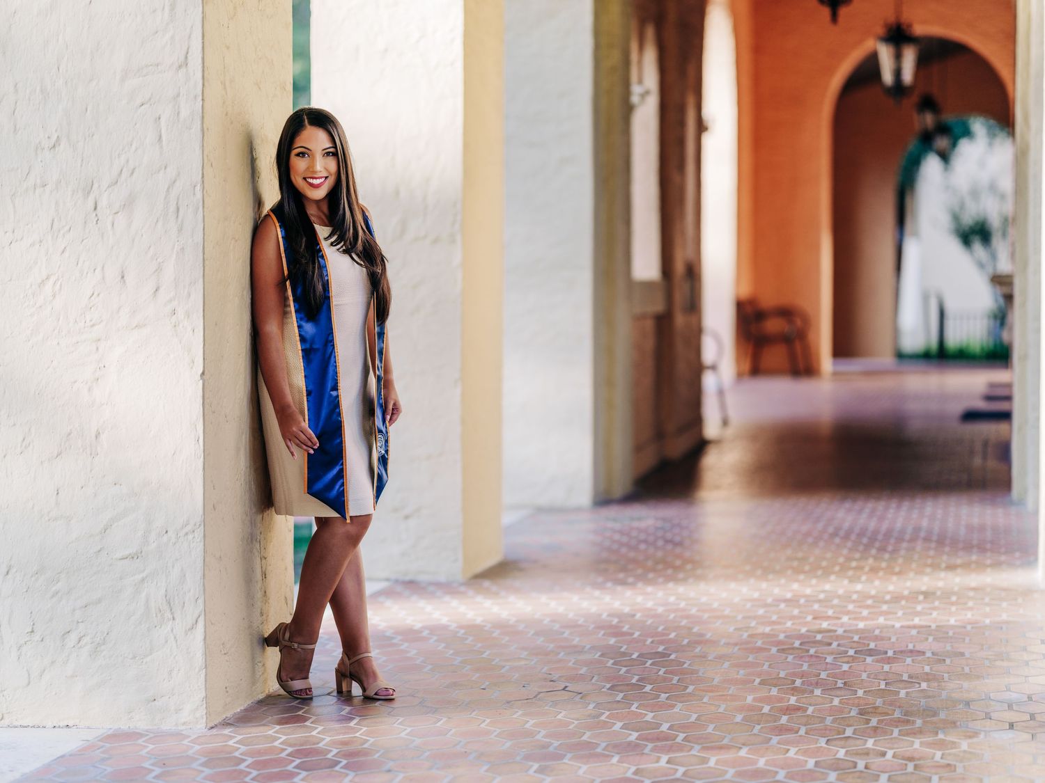 Standing against a beige pillar in a tiled arched hallway wearing a blue dress and long cardigan with heels.