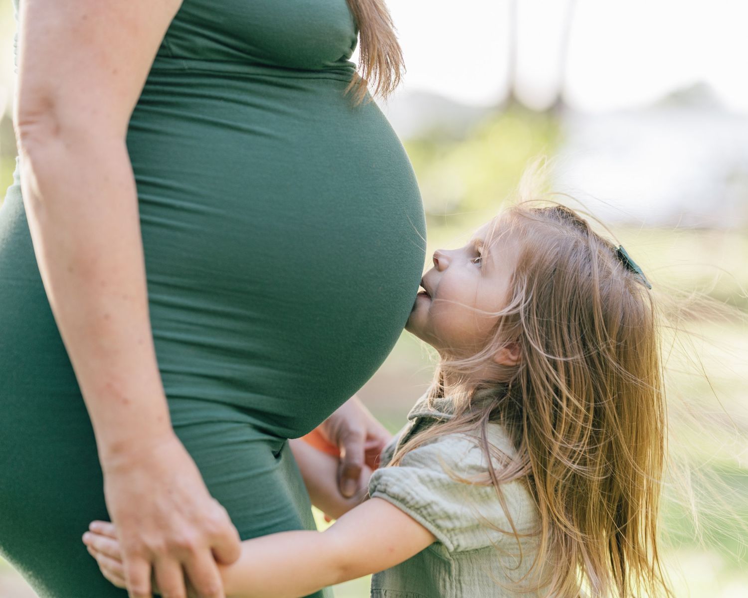 Young child with long hair kisses pregnant belly while standing in outdoor sunlit setting.