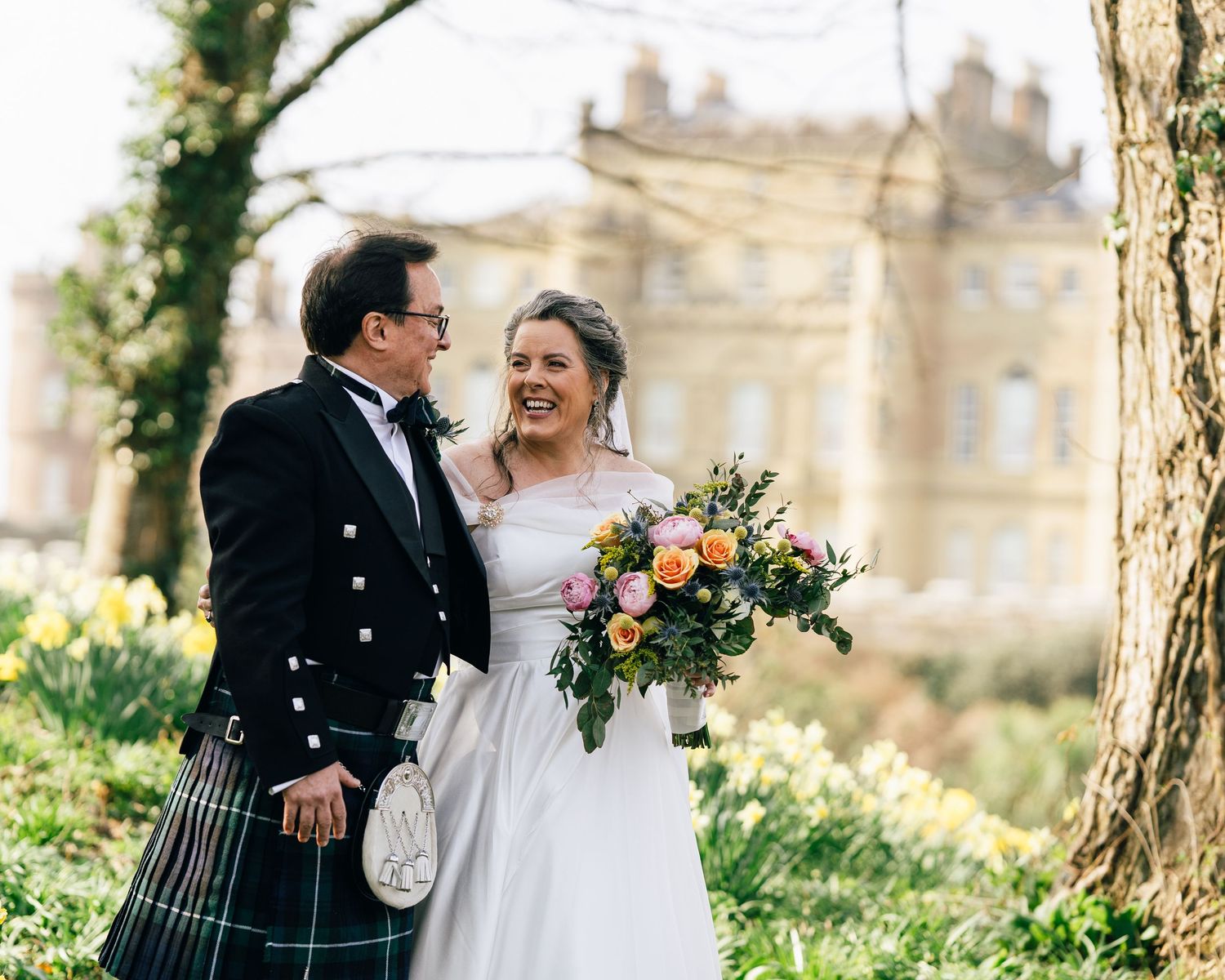 A Scottish wedding couple stands outdoors near the historic Culzean Castle with a colorful bridal bouquet in springtime.