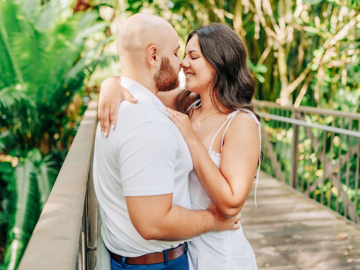 Couple sharing a romantic moment on a bridge surrounded by lush tropical greenery, dressed in white and blue.