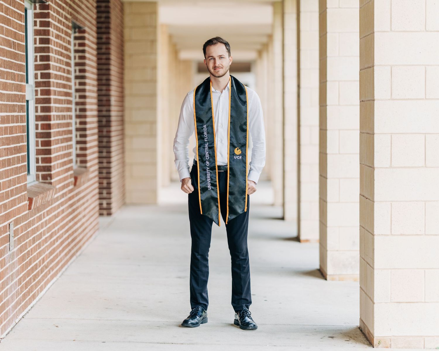 A graduate wearing a dark blue stole and gray blazer stands smiling in a brick colonnade hallway.