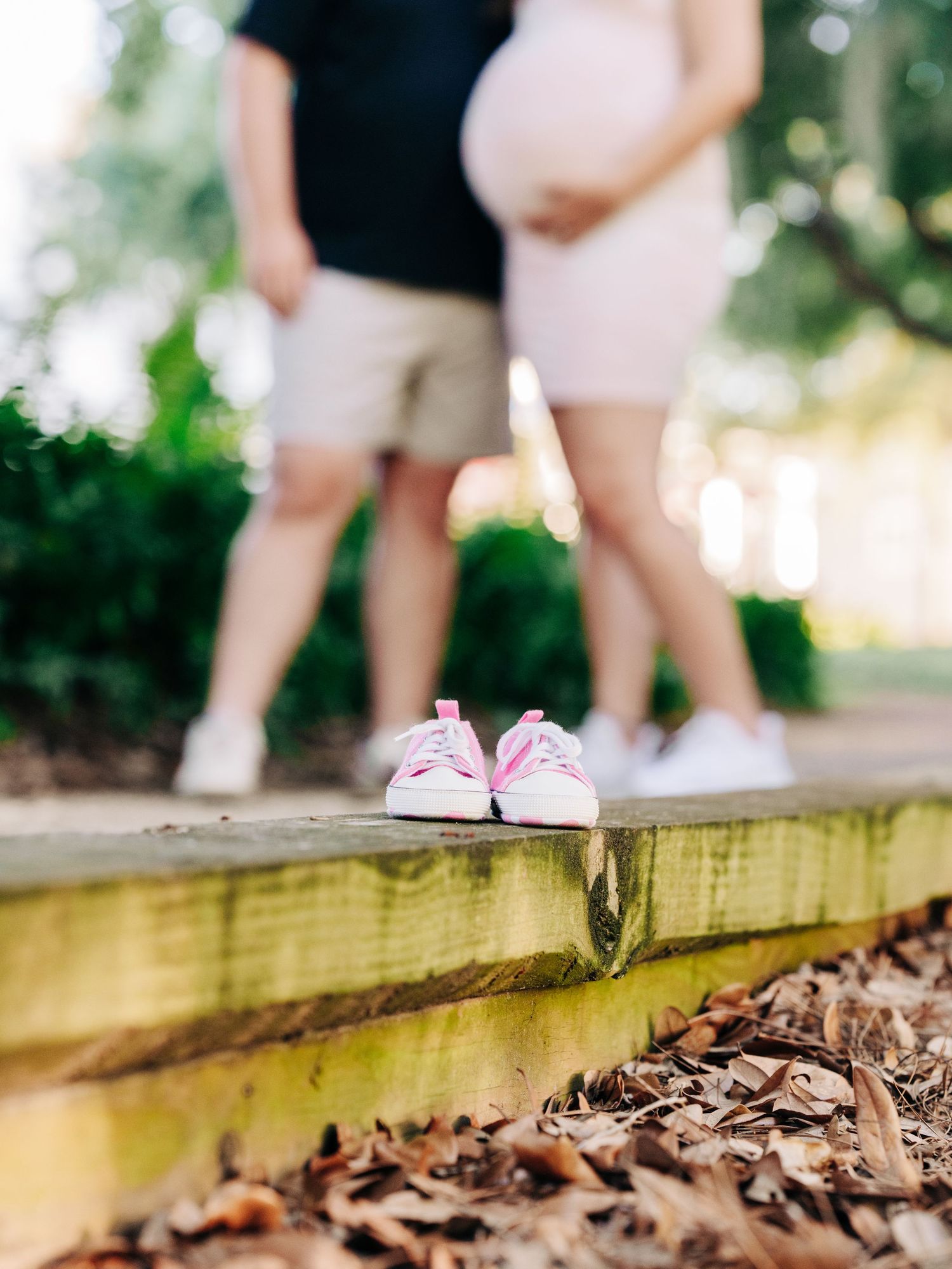 Tiny pink baby shoes sit on a wooden deck with fallen autumn leaves while two people stand in the background.