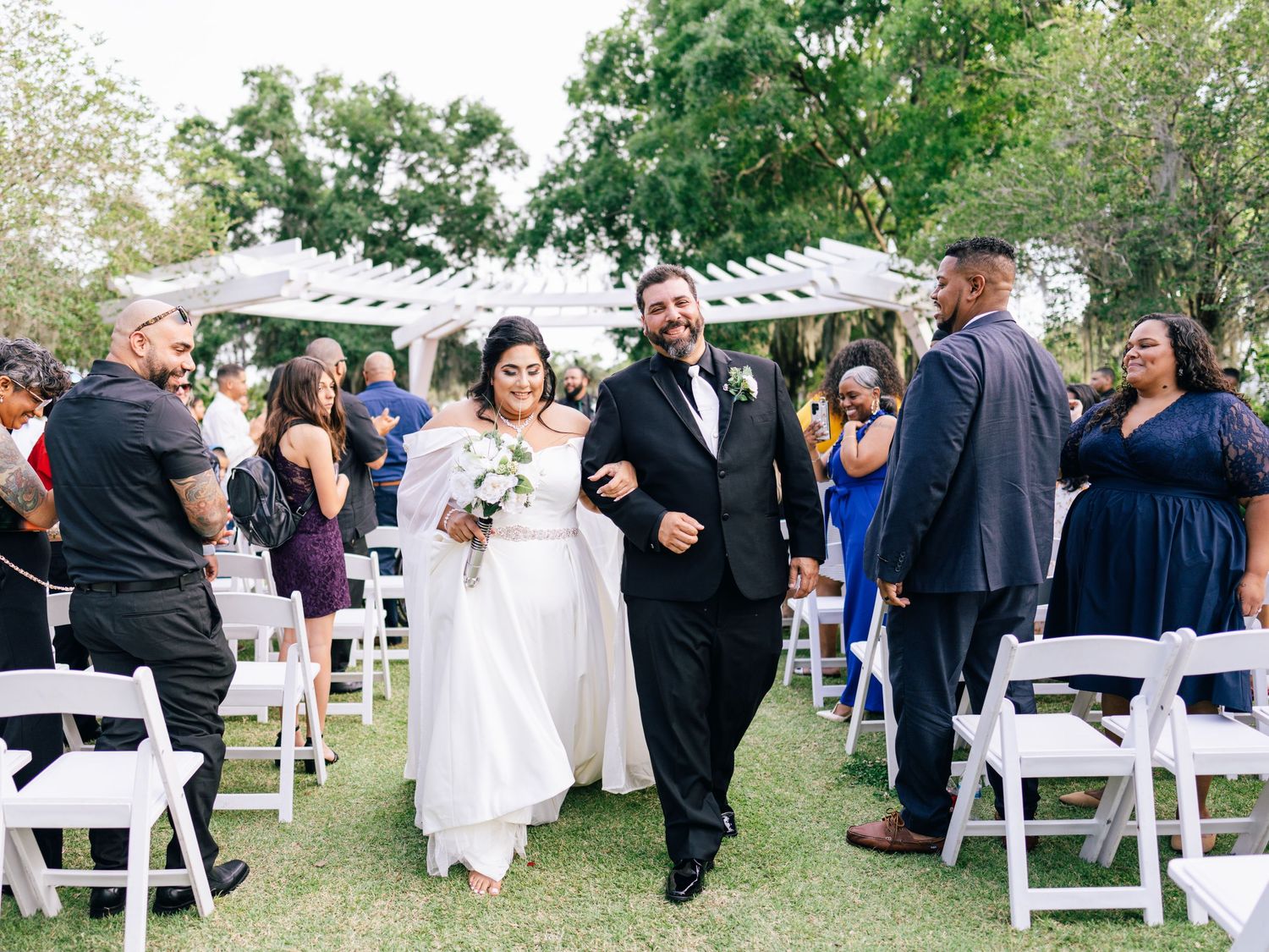 A couple walks down the aisle during an outdoor garden wedding ceremony surrounded by white chairs and guests.
