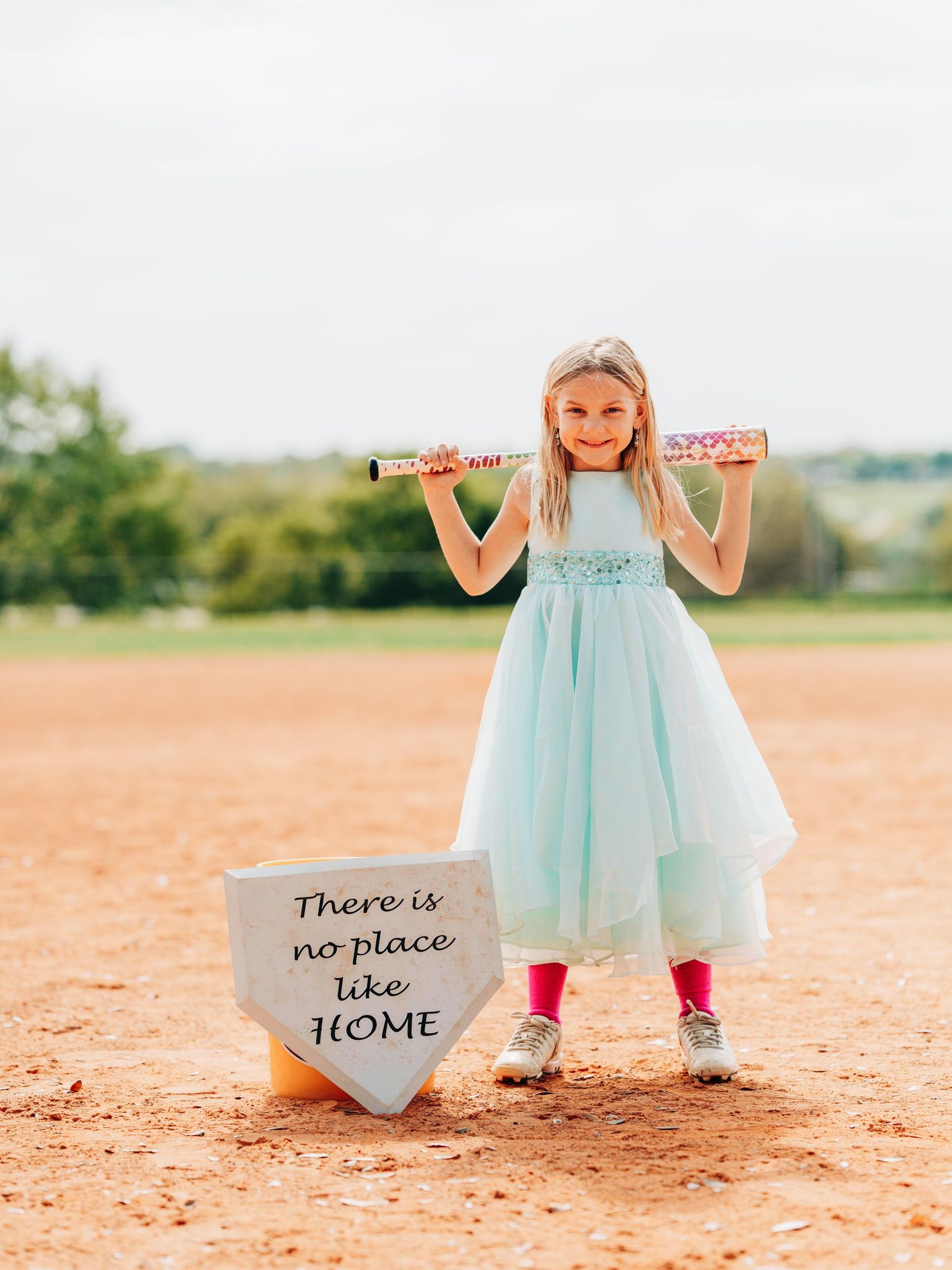 Young athlete on baseball field wearing mint dress and red leggings poses with bat near home plate sign.
