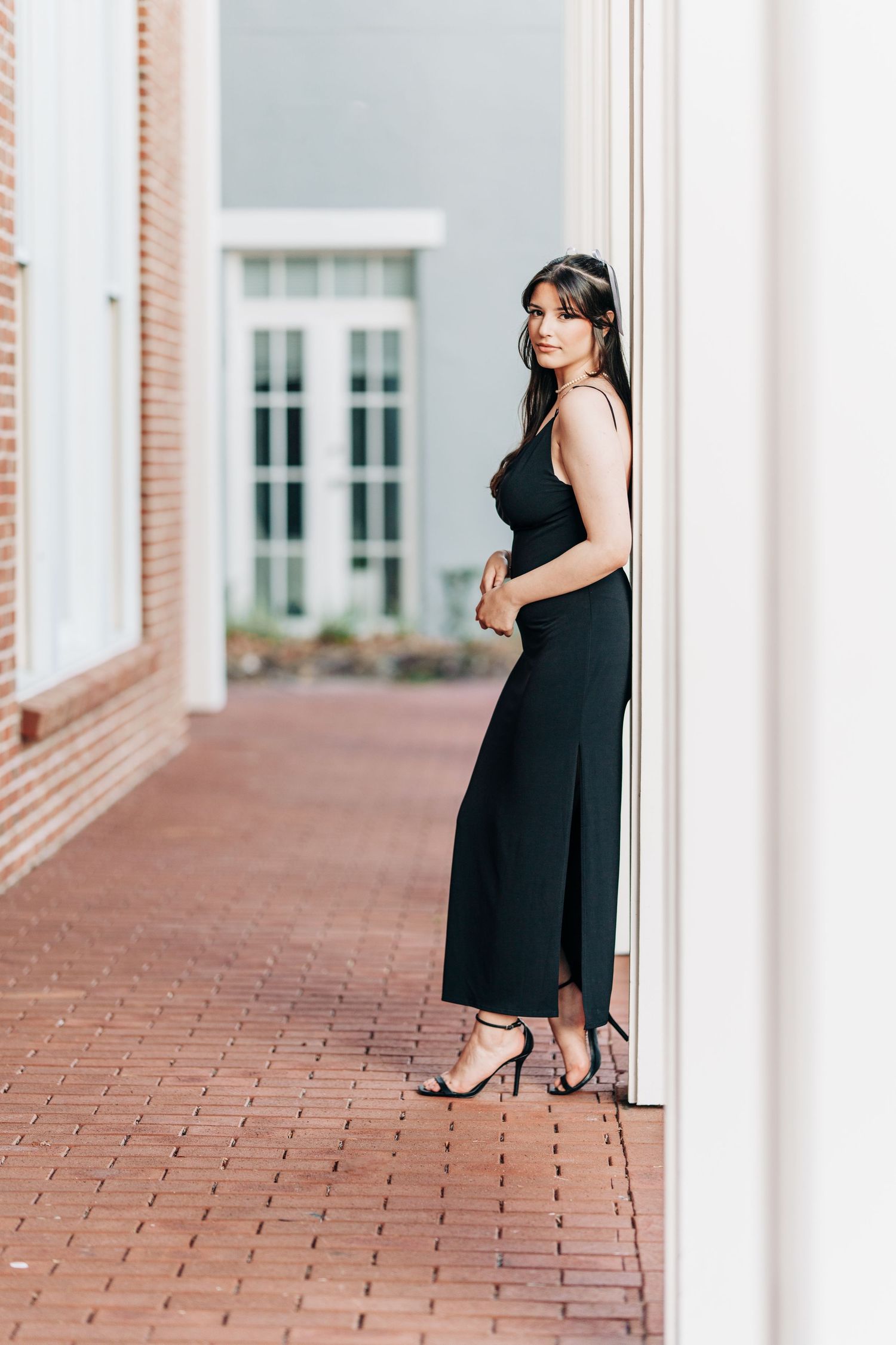 A woman wearing an elegant black maxi dress and heels poses against a white pillar on a brick walkway.