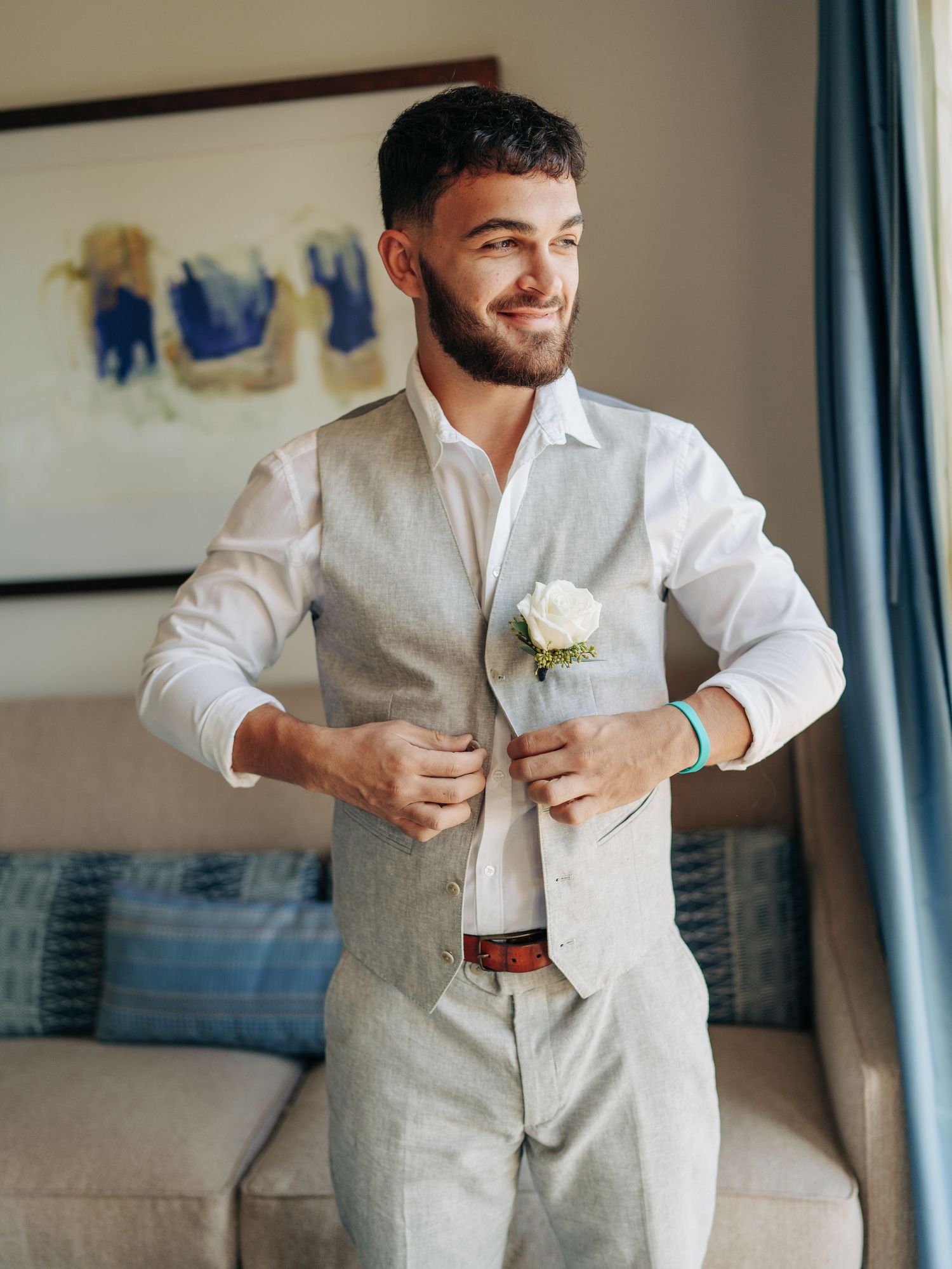 A groom in a light beige suit and white shirt pins a white rose boutonniere while getting ready in a hotel room.