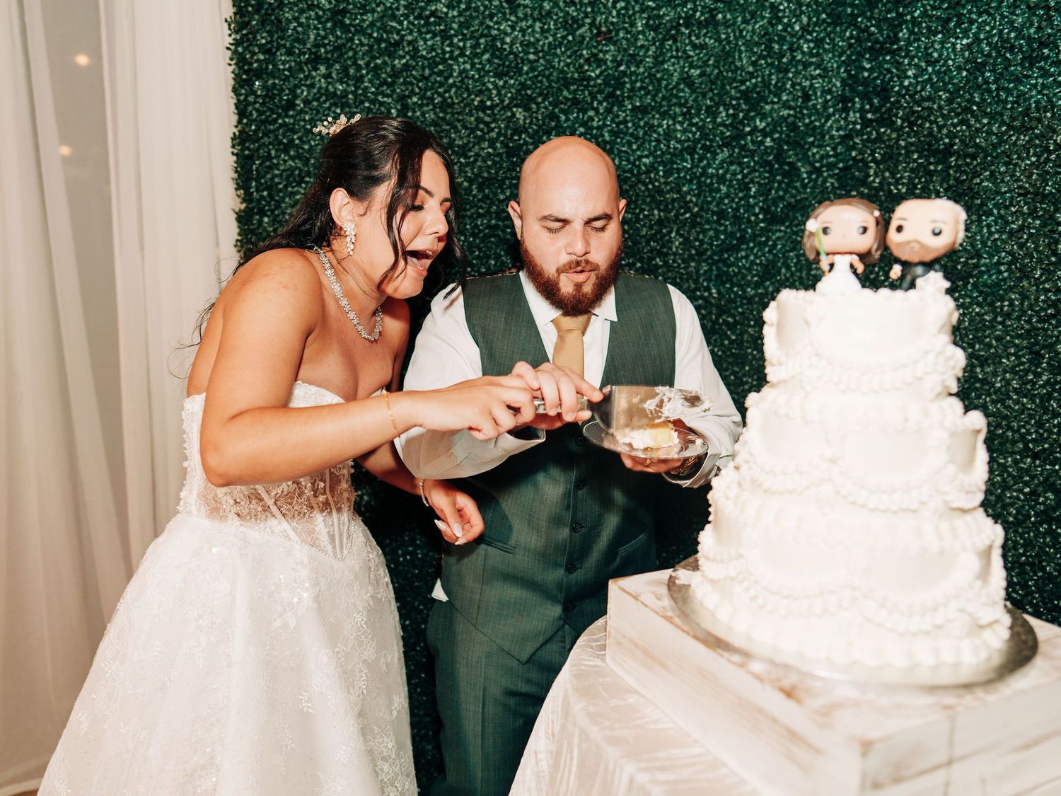 Bride and groom cutting their white tiered wedding cake with custom bobblehead toppers at their reception.