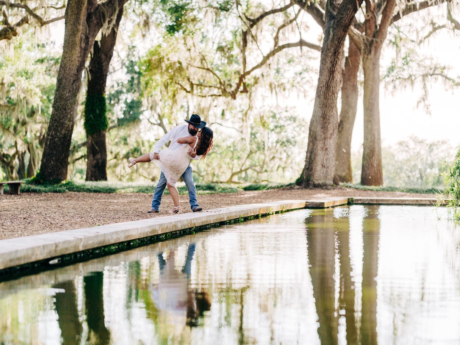 Couple dancing romantically by a reflective pond under moss-draped oak trees in a sunlit park setting.