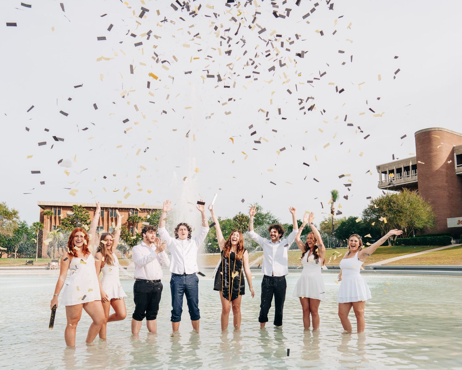 Group of friends in white outfits celebrating with confetti near a fountain outdoors.