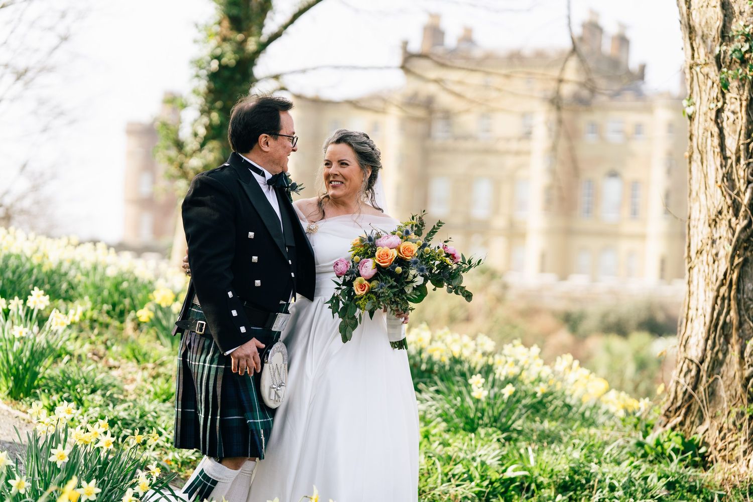 Bride and groom in Scottish kilt holding colorful bouquet among daffodils outside a grand estate in spring.