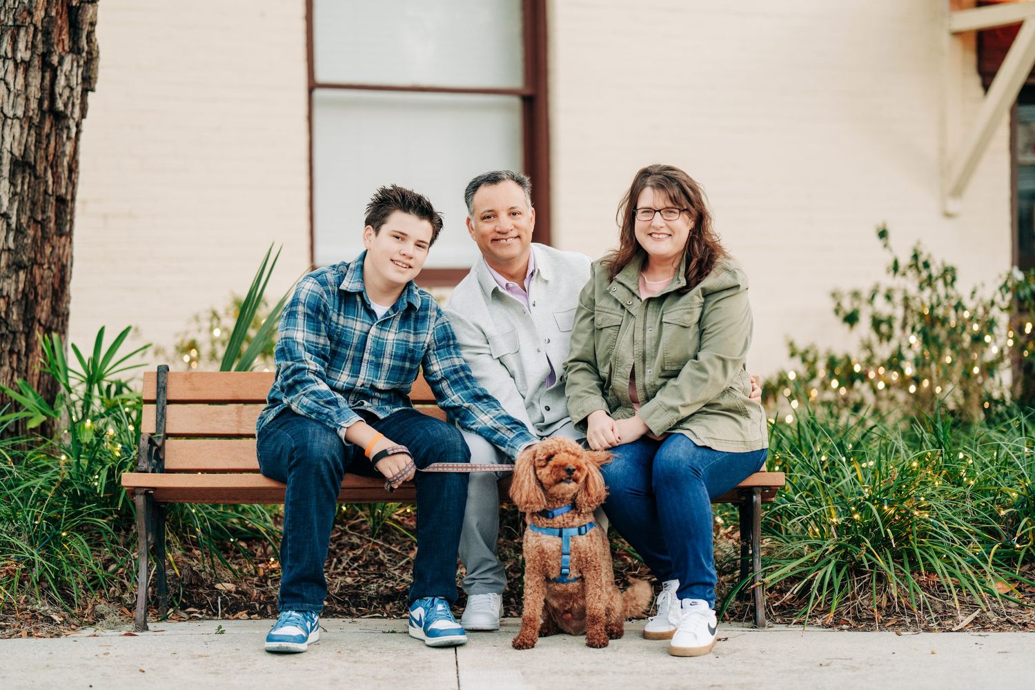 Family of two adults and a child smiling on a bench outdoors with a fluffy red Goldendoodle dog.