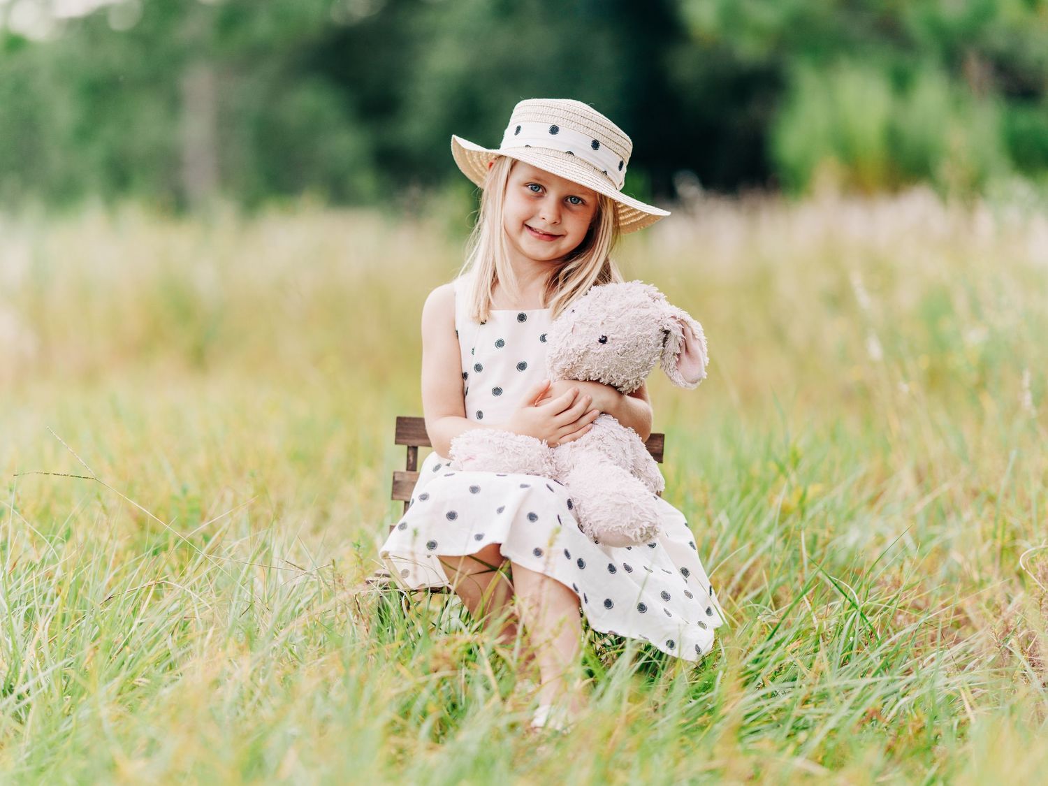 A young child in a polka dot dress and sun hat sits in a grassy summer meadow holding a stuffed bunny.