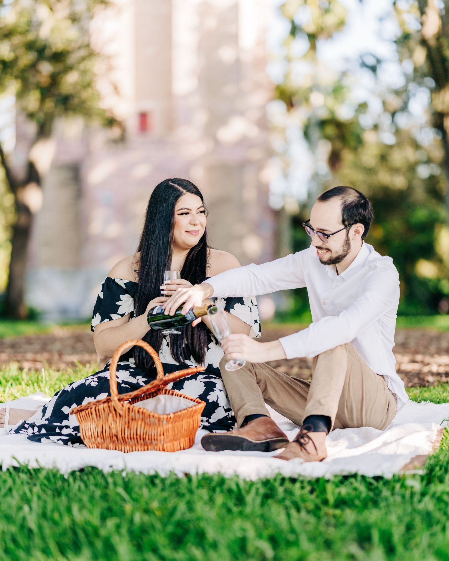 Couple having a romantic picnic on grass with a wicker basket and small dog enjoying a sunny afternoon outdoors.