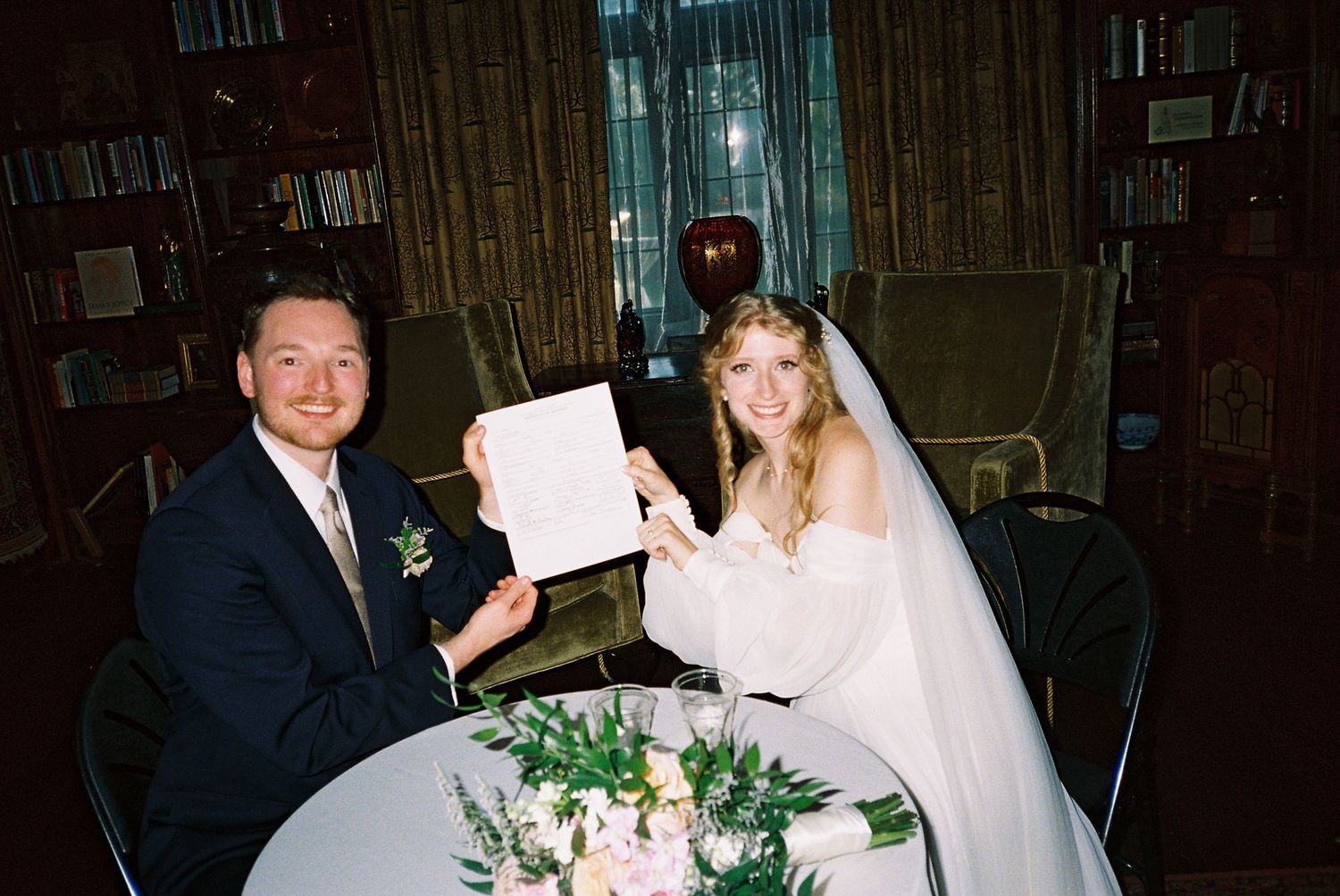 A bride and groom display their marriage certificate while seated at a table with floral arrangements.