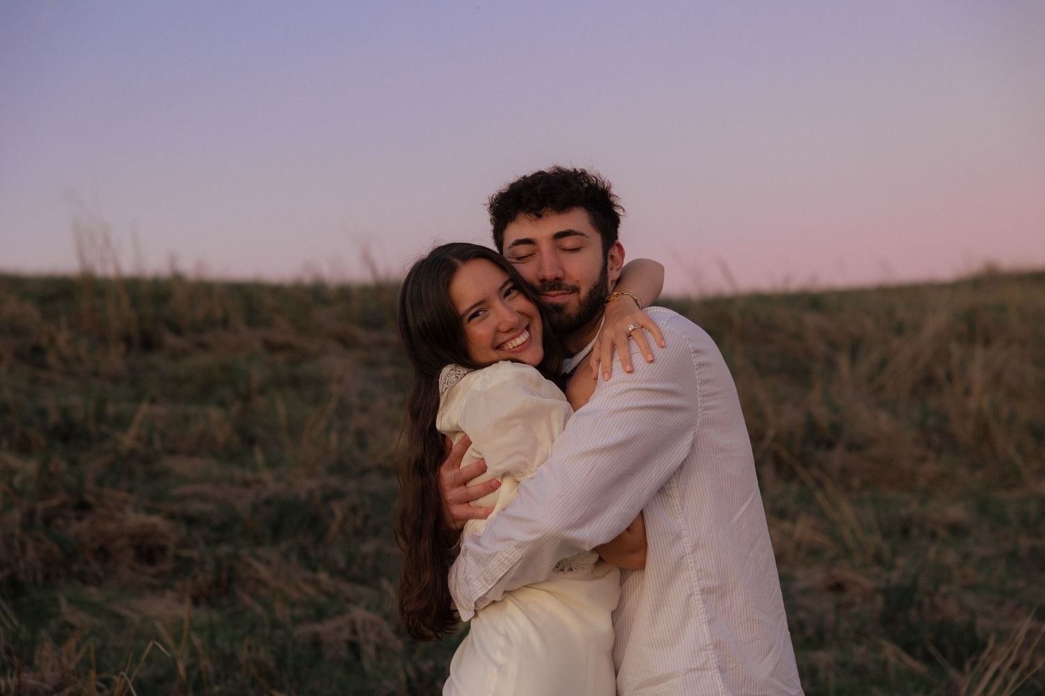 Romantic couple in white outfits embrace during sunset in a grassy field.