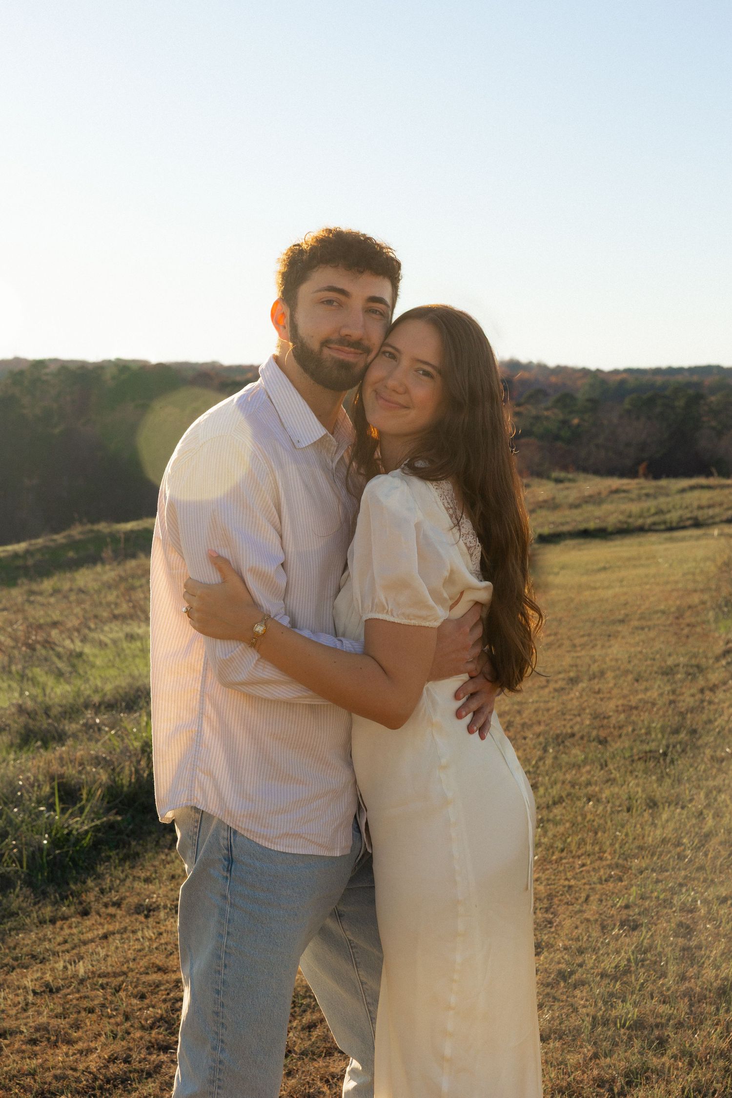 A couple in white outfits poses together on a hilltop during sunset.