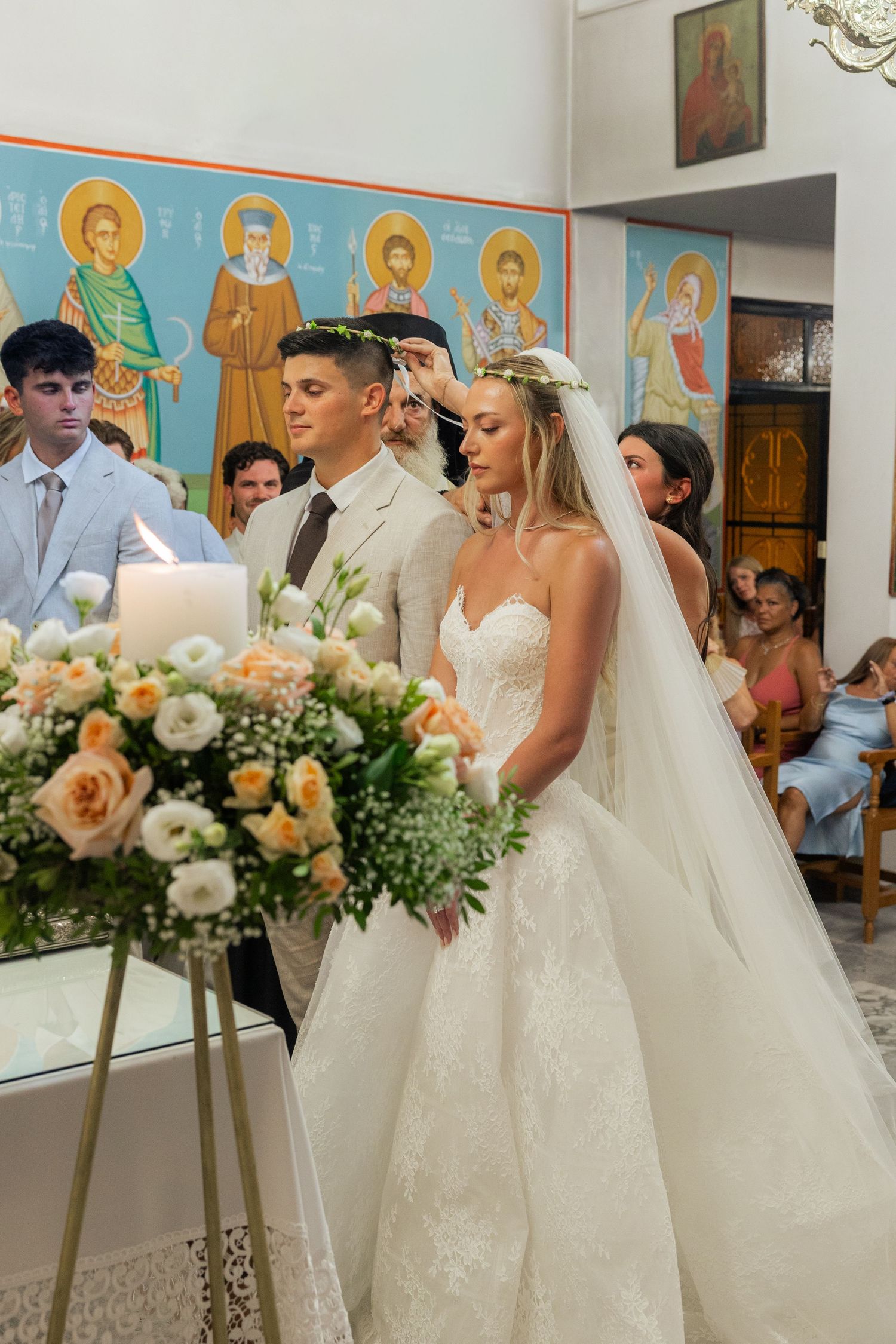 Wedding party members in white dresses and formal attire gather inside a church with religious icons on the walls.