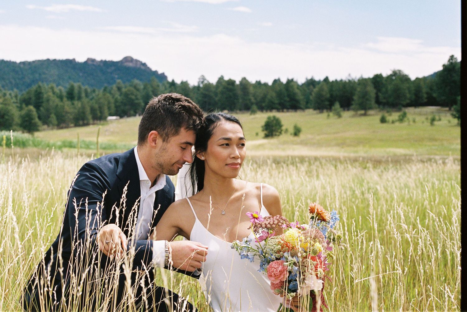 A romantic moment shared between a couple in formal attire amid tall grass in a sunny meadow.