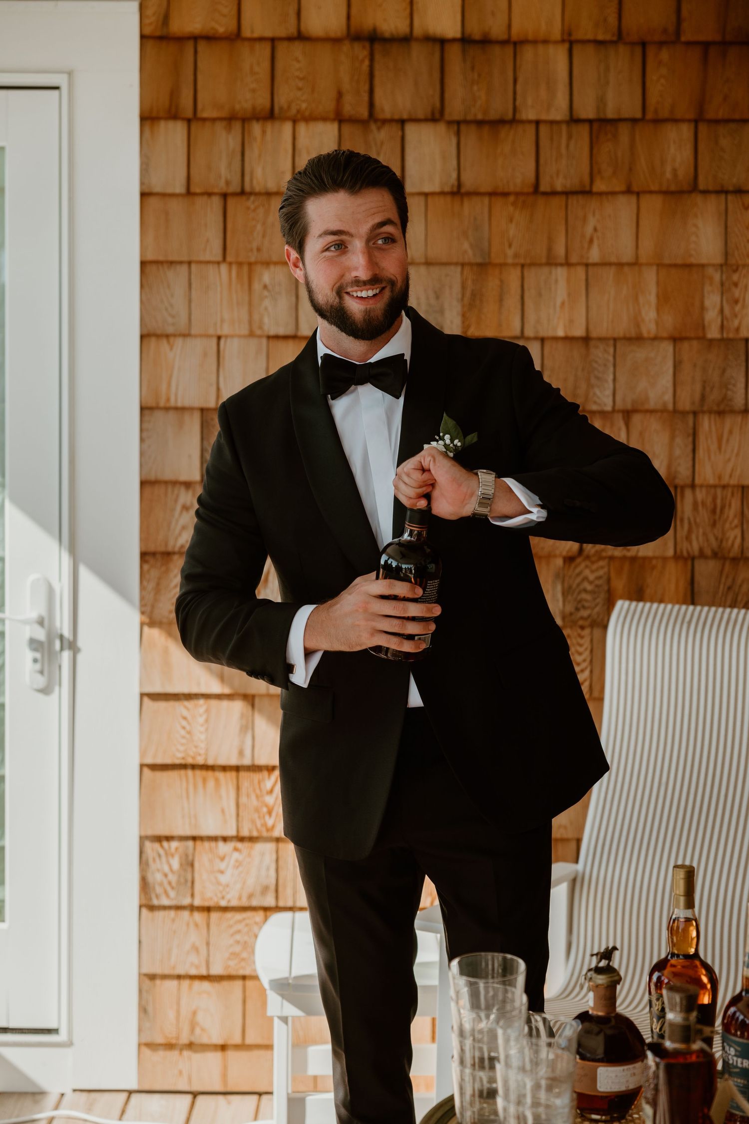 Person in black tuxedo and bowtie holding a drink bottle against cedar shake siding.