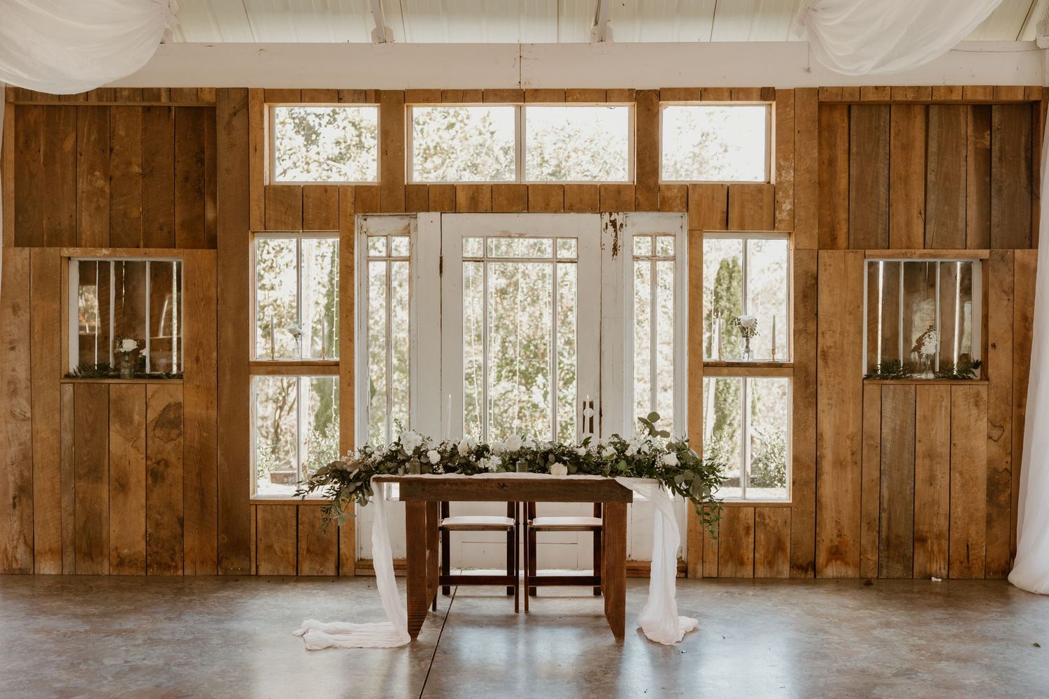 Rustic wooden wedding venue interior with draped table and floral arrangements by large windows.