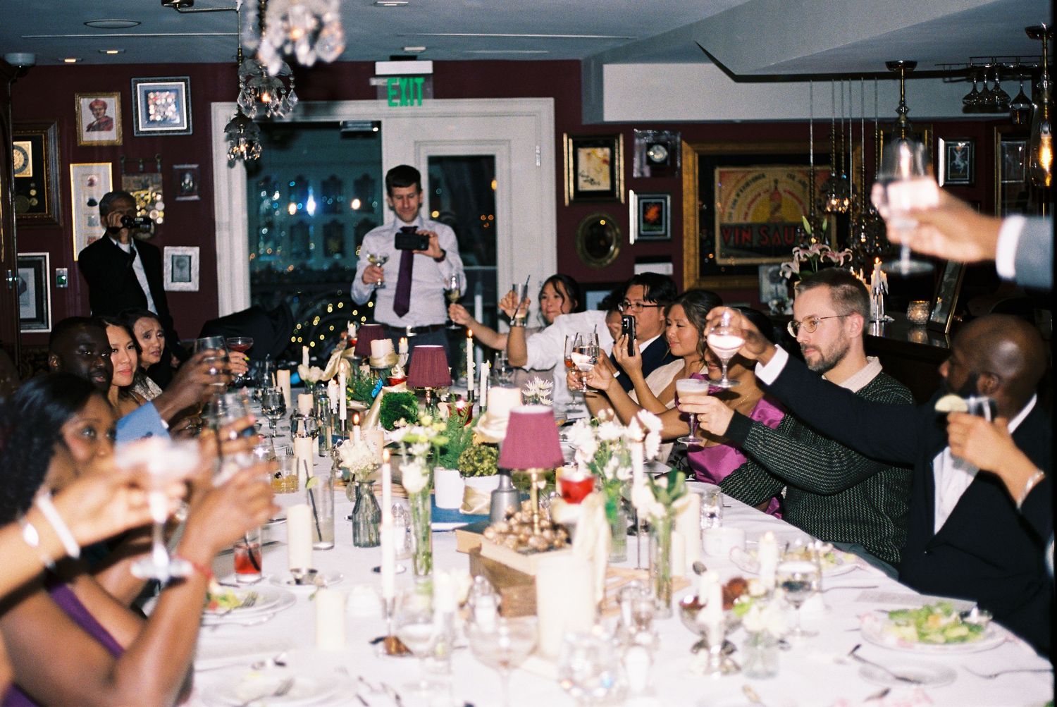 Guests gathered around elegantly set dining tables at an evening reception with crystal chandeliers overhead.